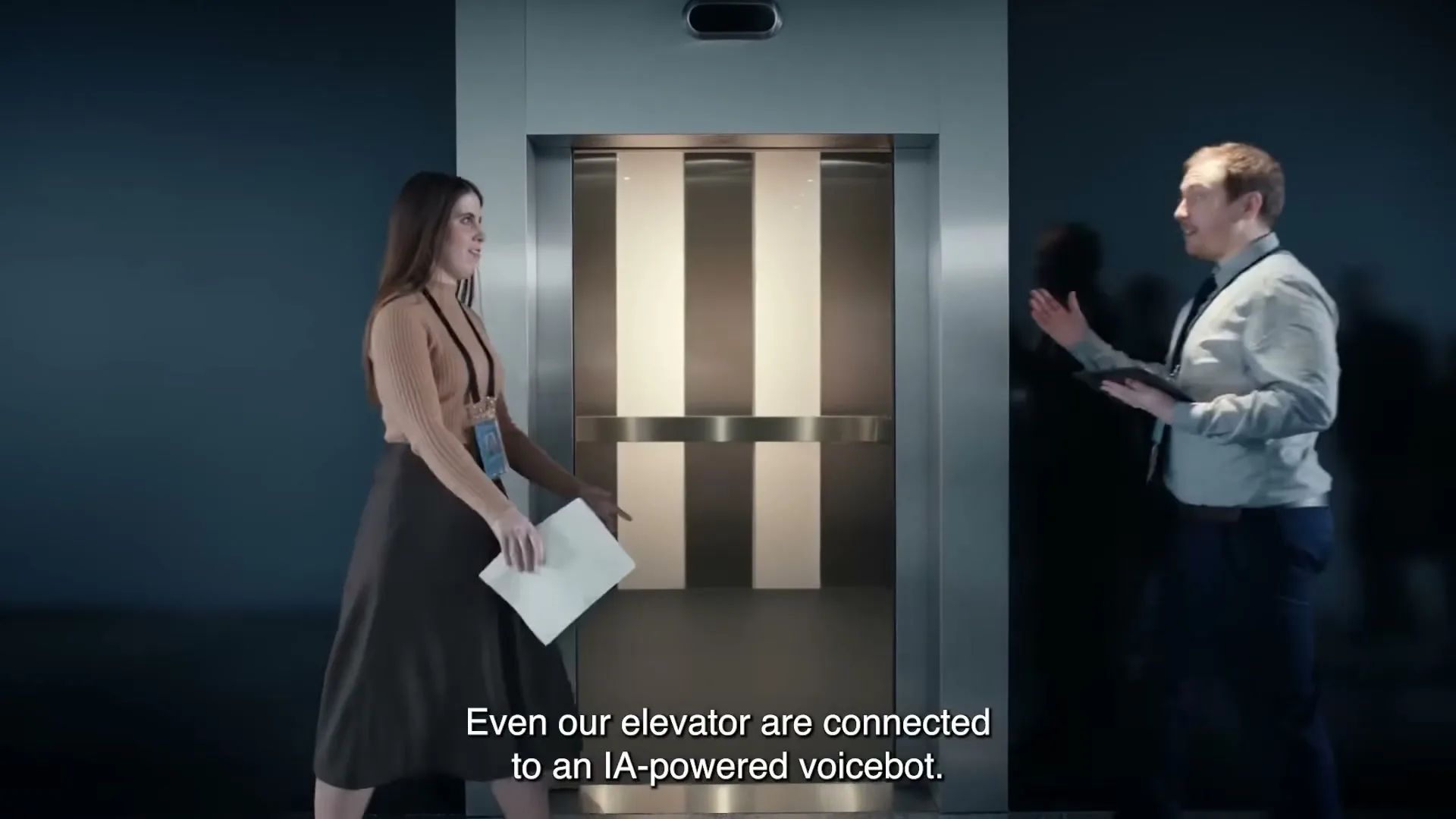 A woman in a brown top and dark skirt approaches an elevator, which opens to reveal a man and another woman entering, in an office building.