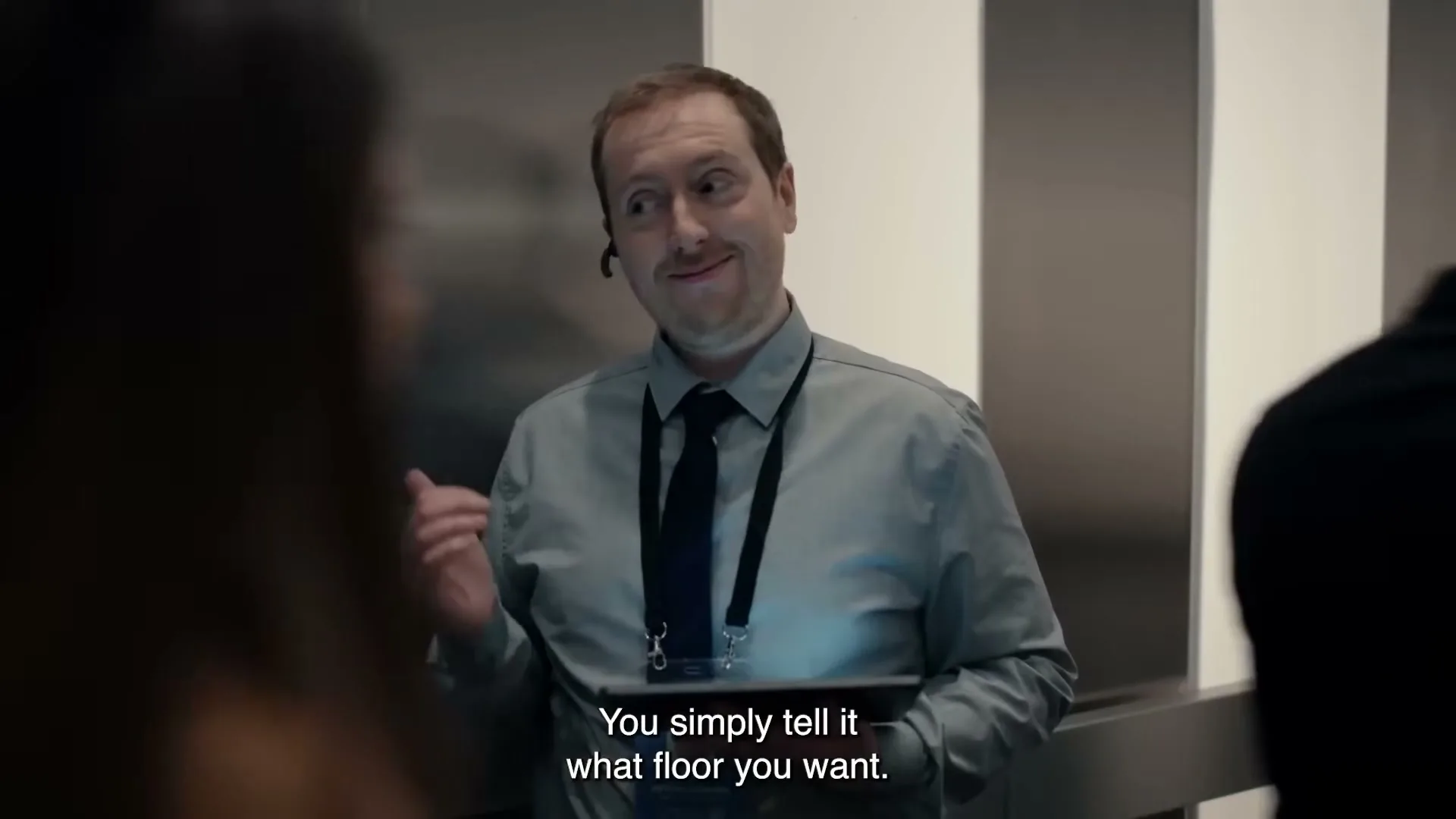 A man in a grey shirt and tie, wearing a headset, holds a tablet and smiles at a woman in an elevator, with a digital screen showing weather and a microphone icon on the wall.