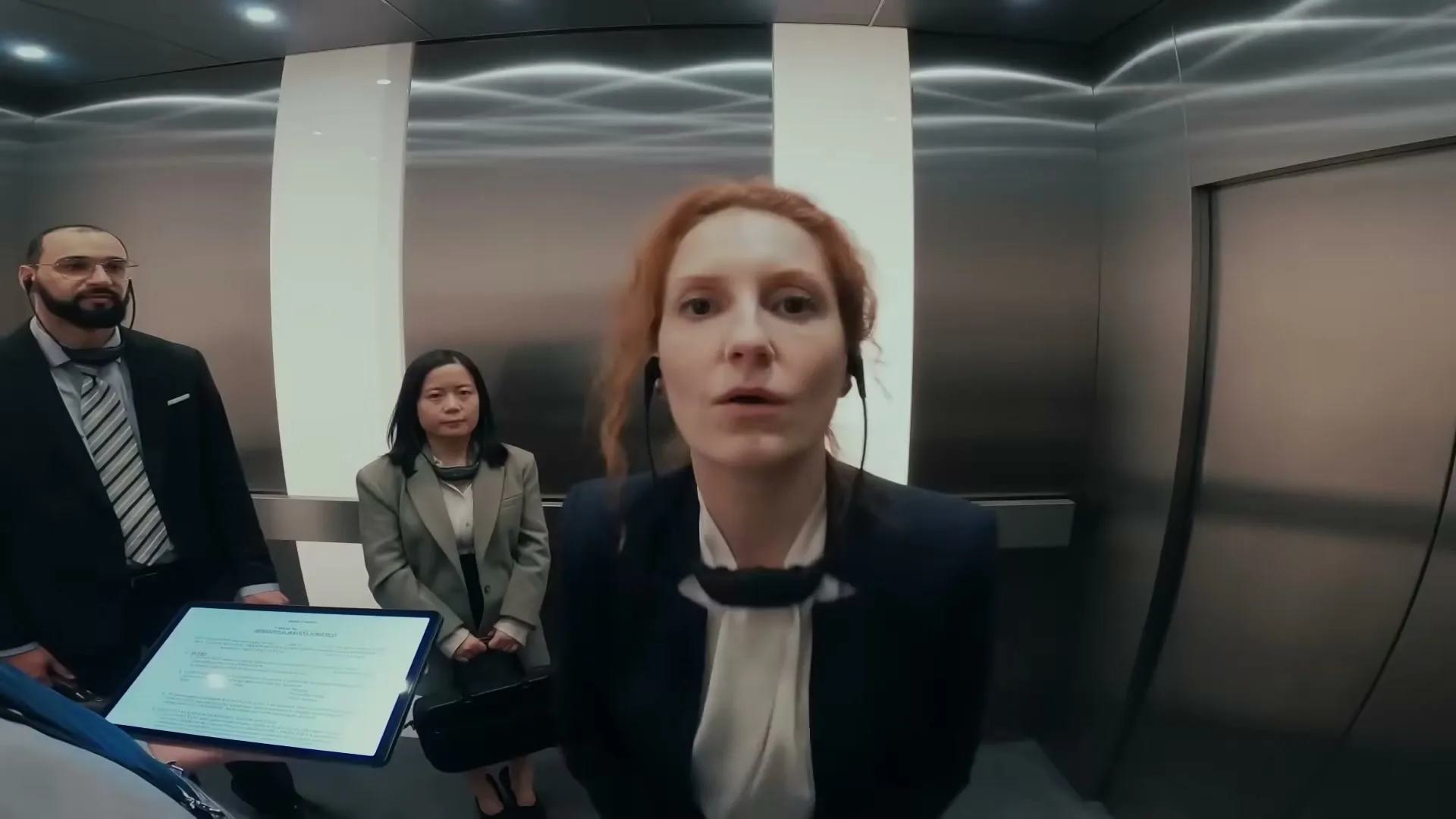A red-haired woman in a business suit looks directly at the camera, standing in an elevator with two other people.