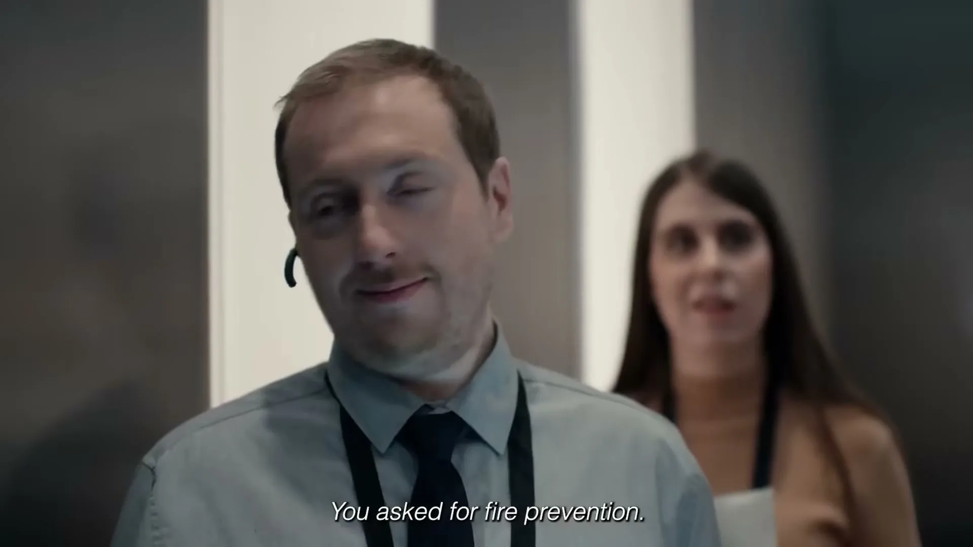 A man with a headset and a woman behind him are in an elevator, as the man looks around with a slight smile.