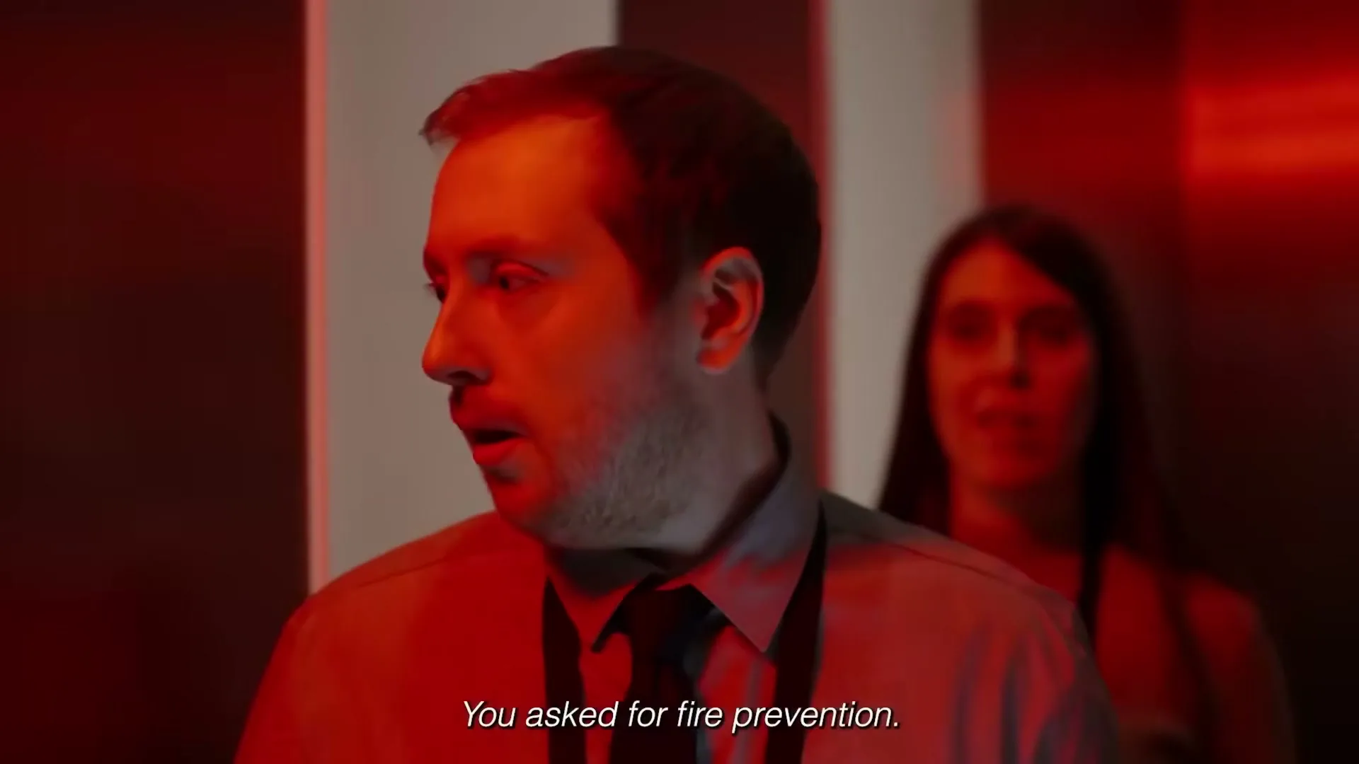 A man in a shirt and tie looks surprised as a woman stands behind him in an elevator, with red emergency lighting.