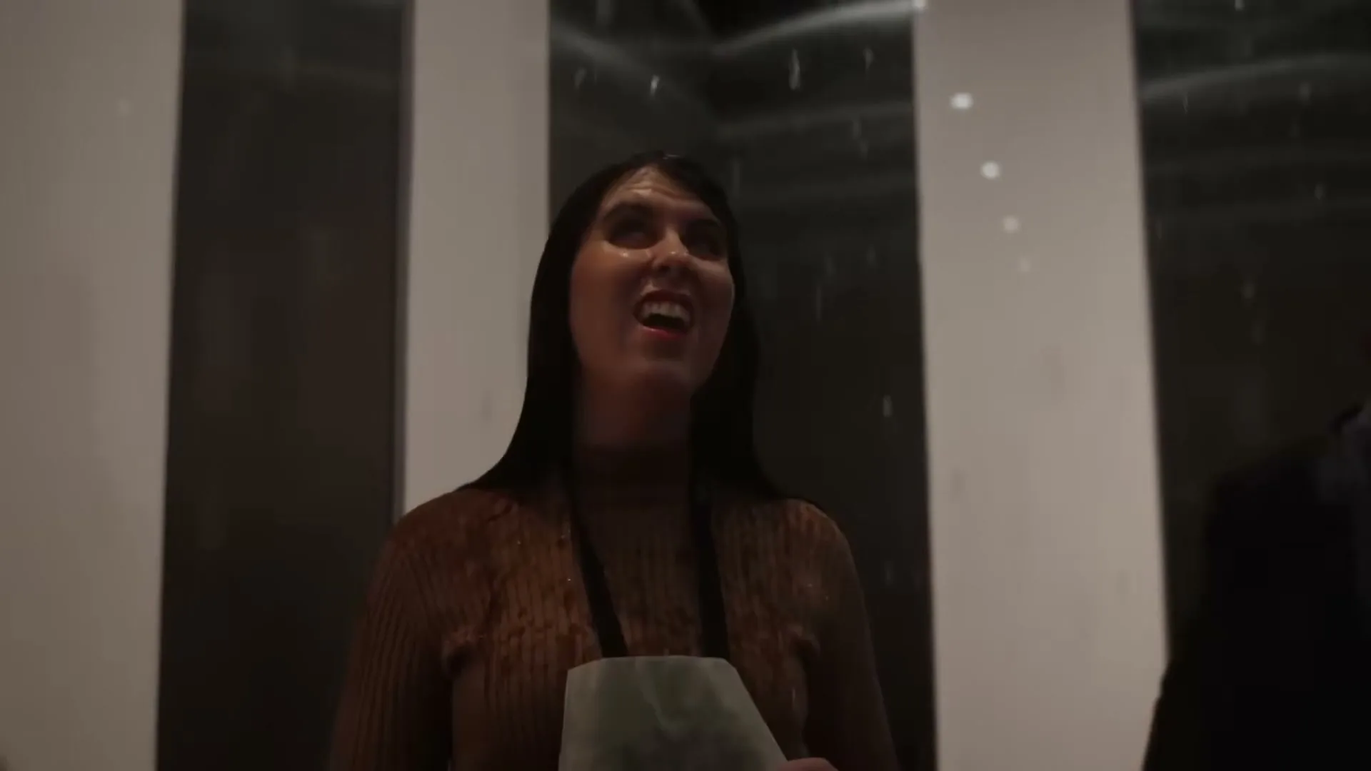 A woman with long dark hair, wearing a brown top, looks up in an elevator as water drips from the ceiling, illuminated by red light.