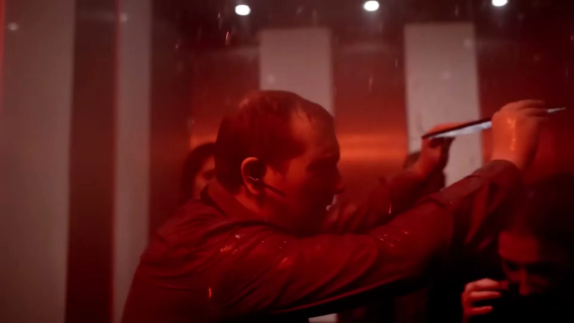 A man in a suit, with an earpiece, holds up a tablet in a dark, red-lit, water-filled elevator with other people.