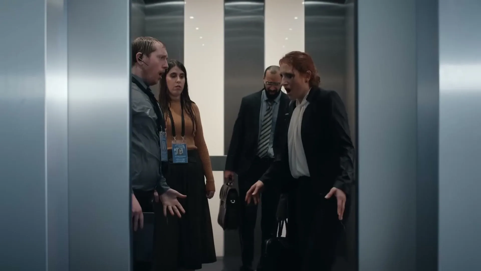 A group of four office workers, two men and two women, are seen inside an elevator with surprised expressions as the doors open.