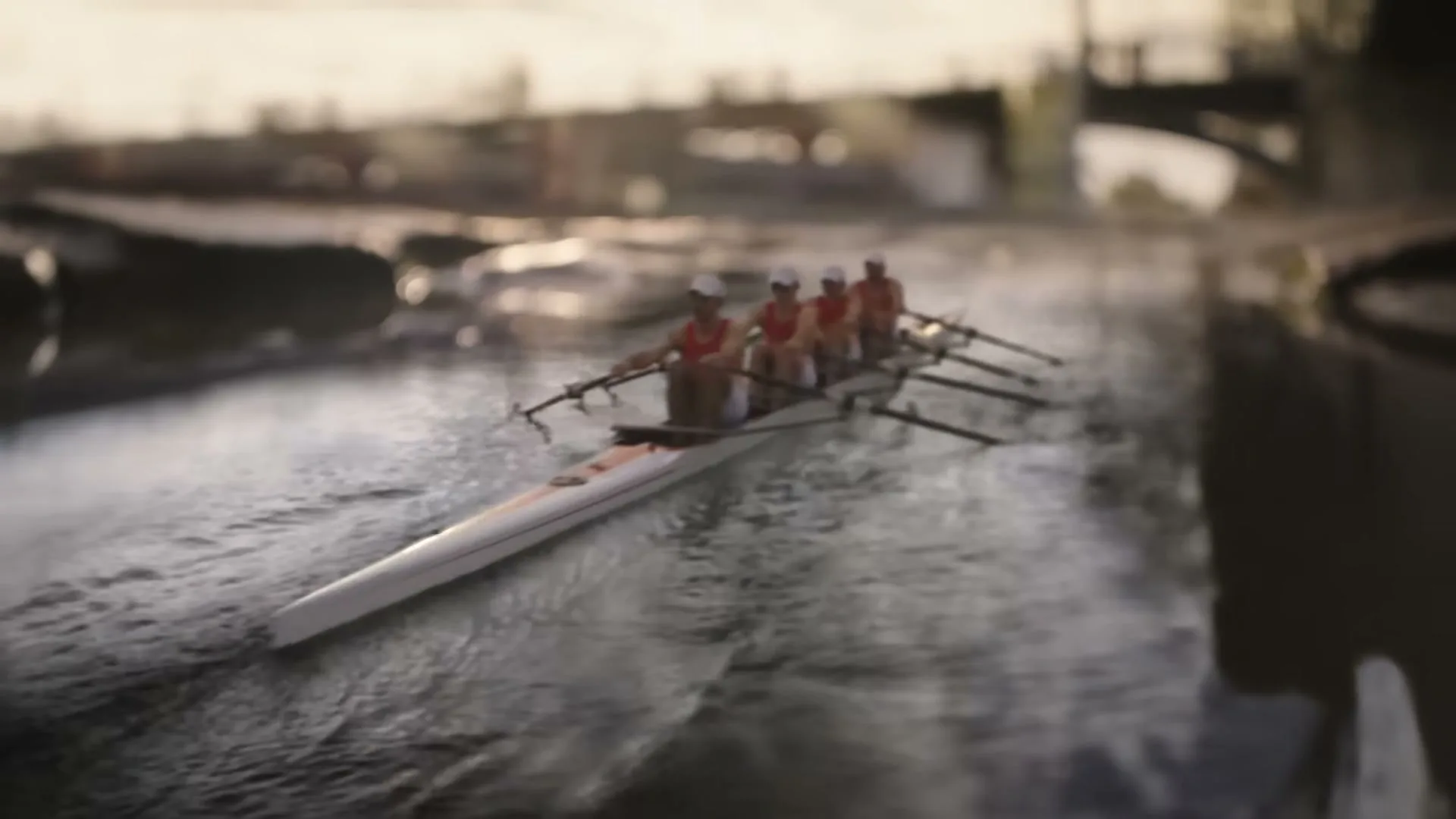 Four rowers in red and white uniforms paddle a long, narrow boat on a river, with a bridge in the blurry background during golden hour.