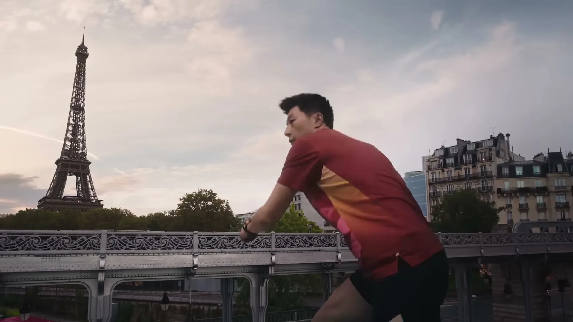 A male table tennis player in a red shirt and black shorts plays table tennis on a bridge in Paris, with the Eiffel Tower in the background.