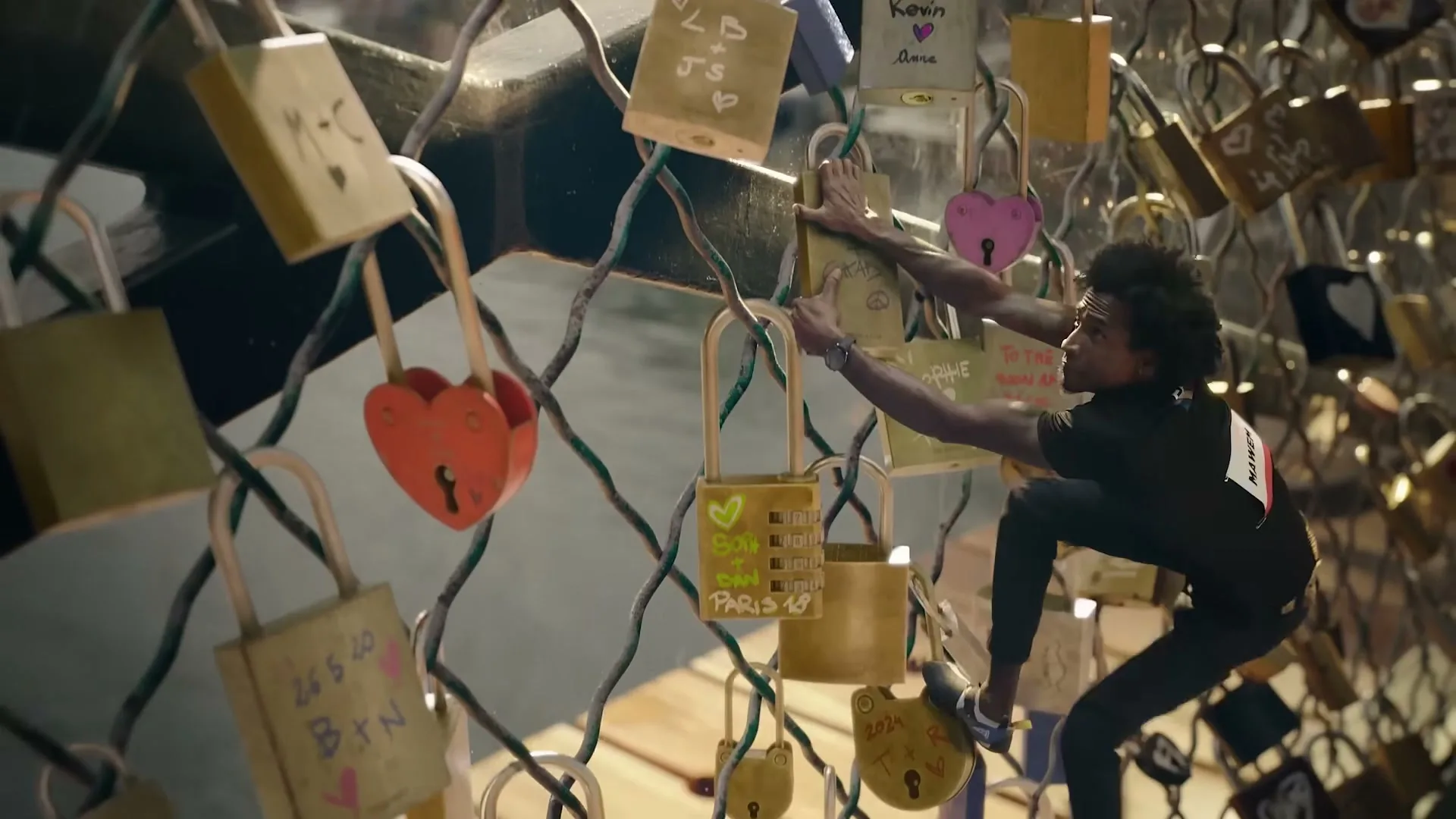 A male athlete in a black shirt and pants climbs a fence covered in love locks, with a river visible in the background.