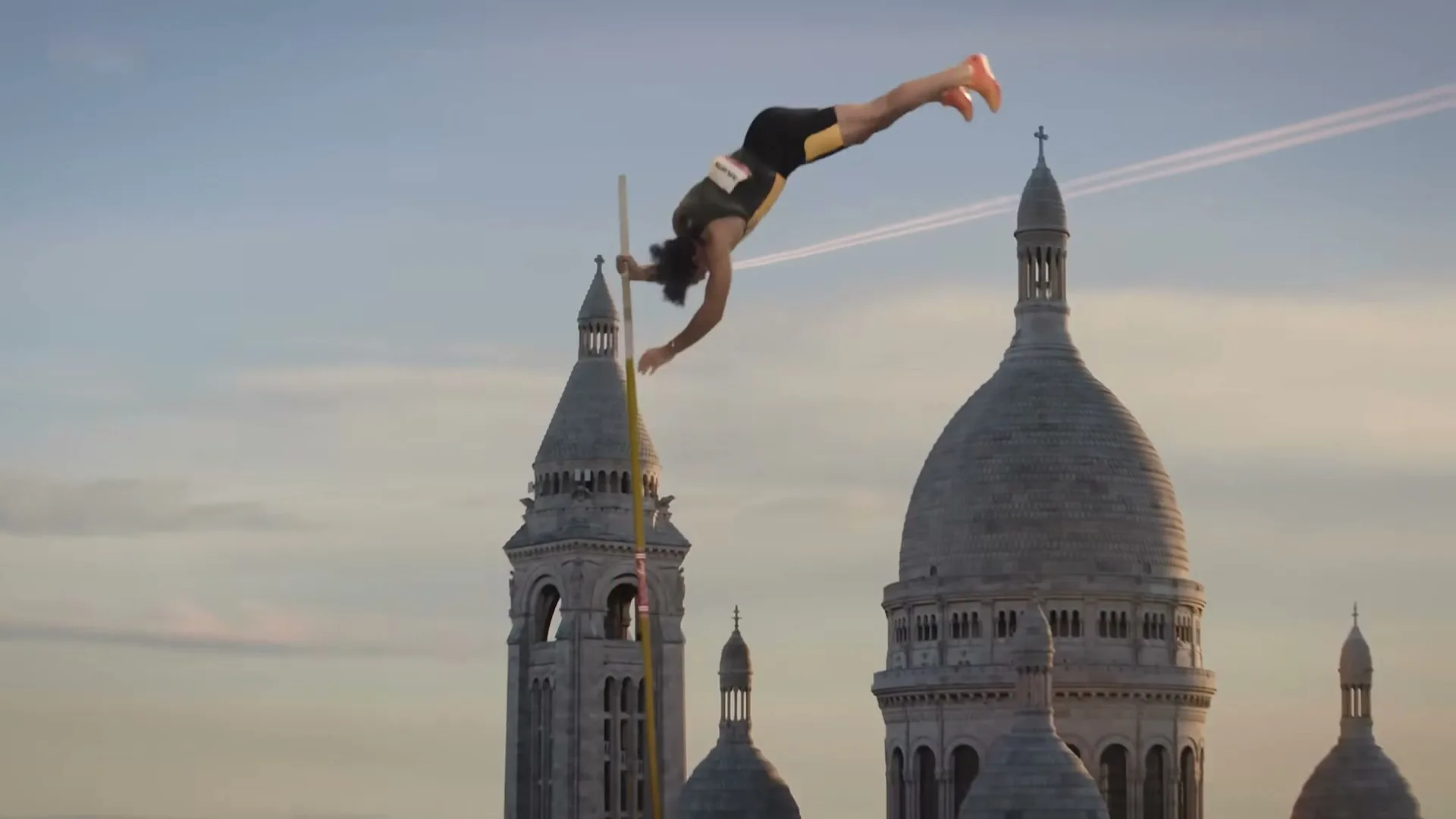 A male pole vaulter in athletic wear soars over the Sacré-Cœur Basilica in Paris, France, at golden hour.