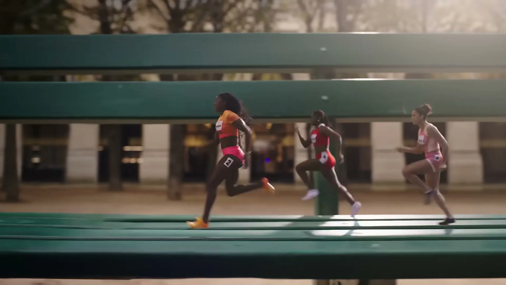Three female athletes in athletic wear run on a track in a park, with a green bench in the foreground.
