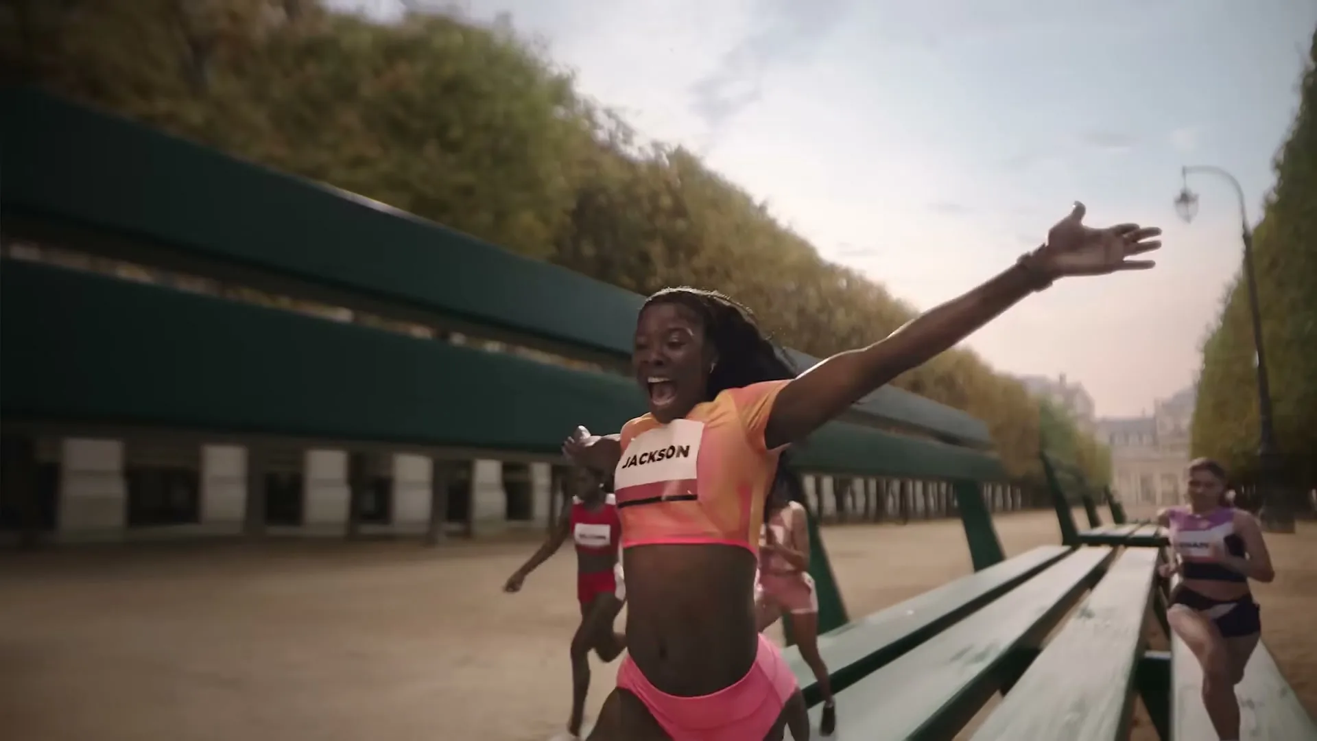 Elaine Thompson-Herah, a female athlete, runs with arms outstretched and a joyful expression in a park with benches and trees.