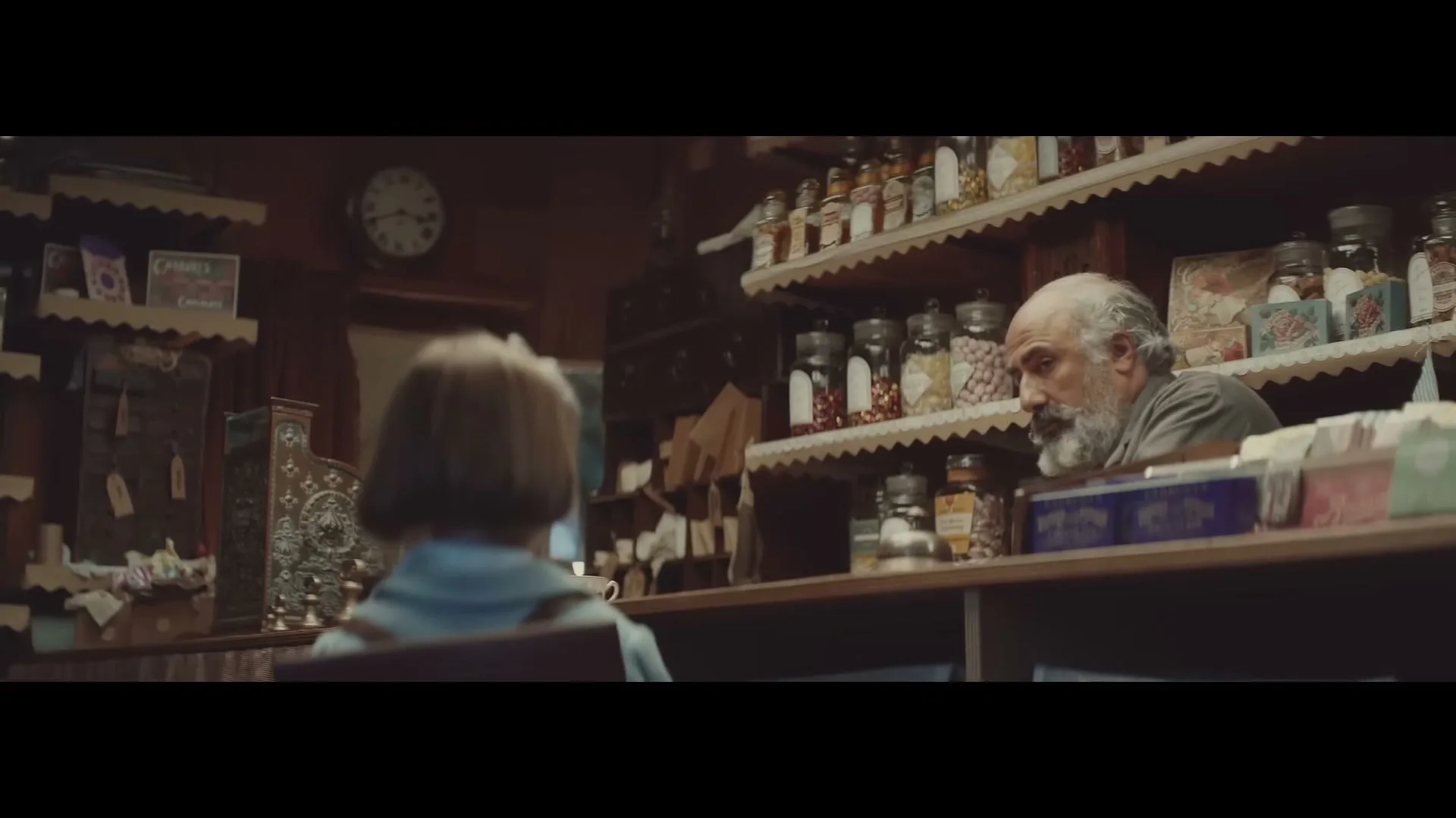A young girl with a bob haircut sits at a counter, looking at an older man with a beard behind the counter in a rustic shop.