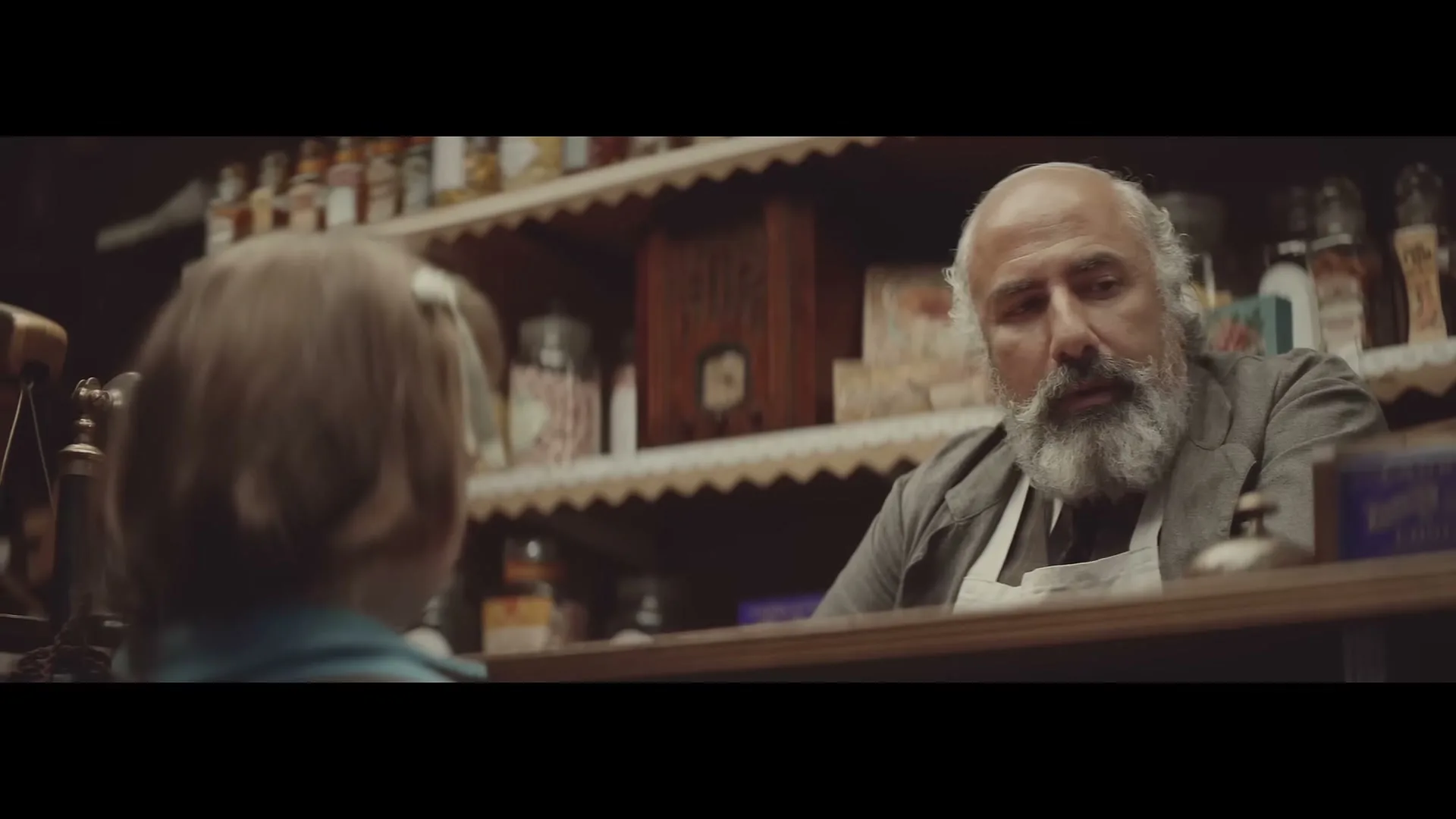 A young girl with brown hair stands in a sweet shop, looking up at a kind-faced, bearded shopkeeper behind the counter.