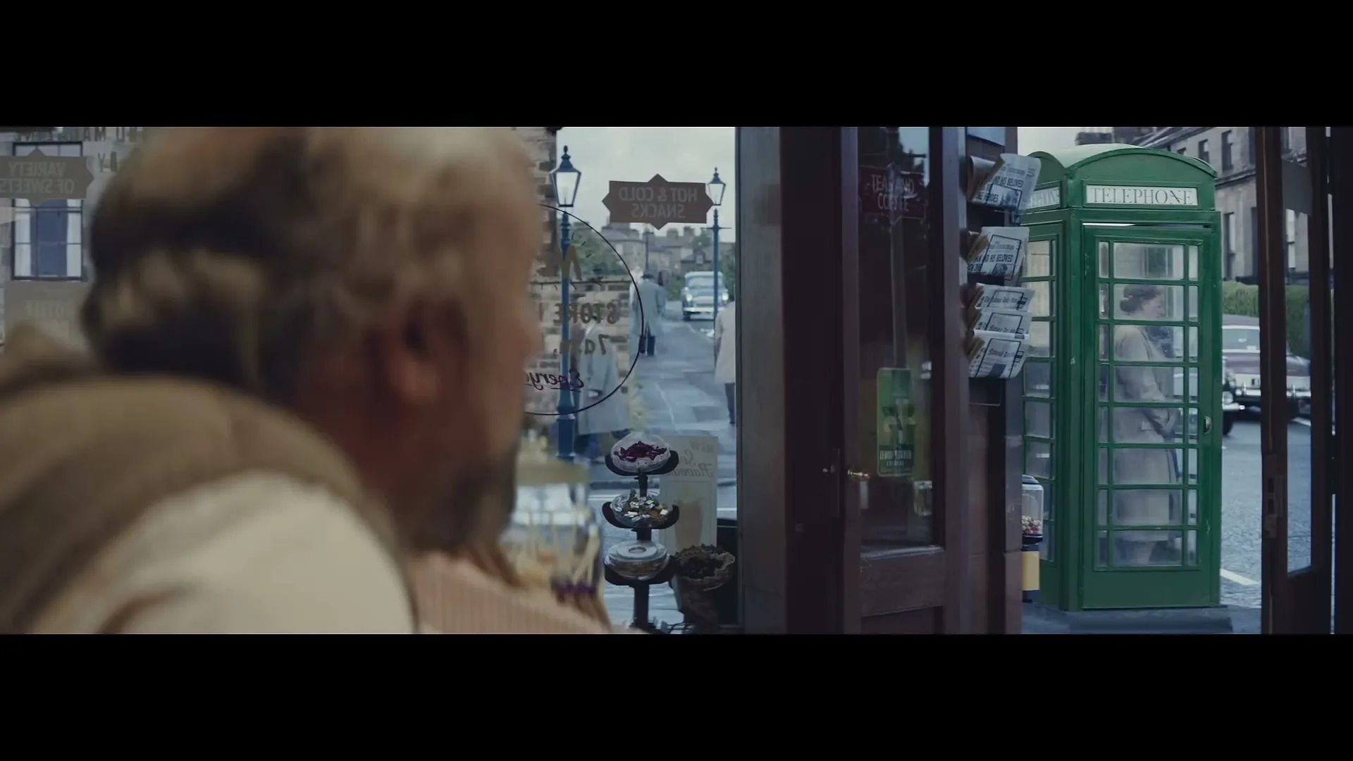 A man with a beard looks out a shop window at a woman in a phone booth on a street.