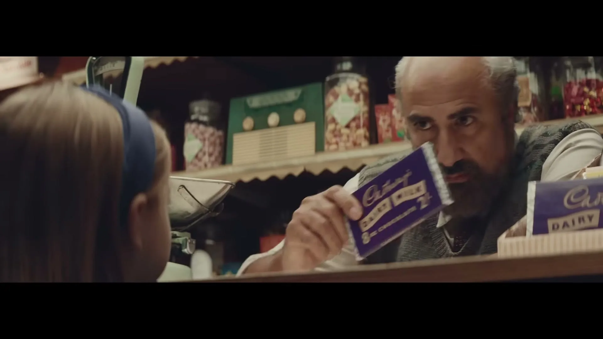 A shopkeeper with a beard holds up a Cadbury Dairy Milk chocolate bar to a young girl in a sweet shop.