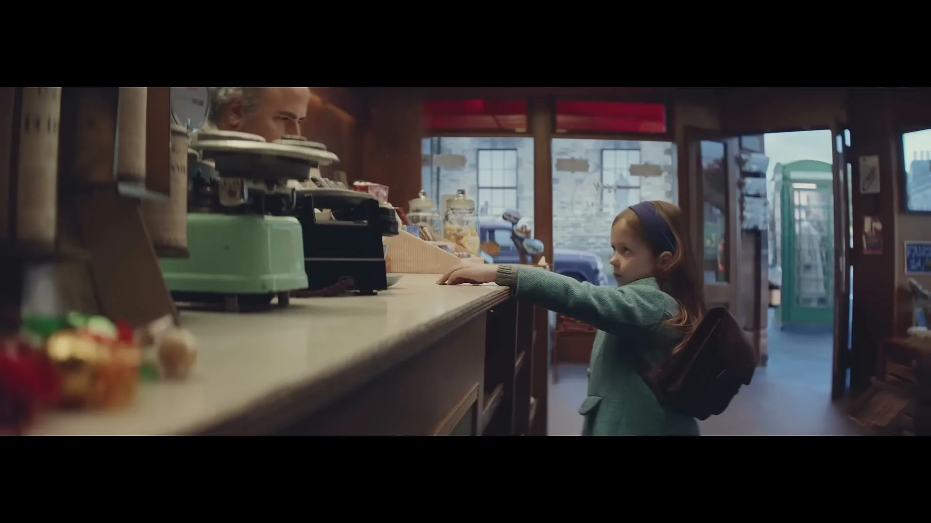 A young girl in a blue coat reaches across a counter towards a shopkeeper in a vintage sweet shop, with an old-fashioned street visible outside.