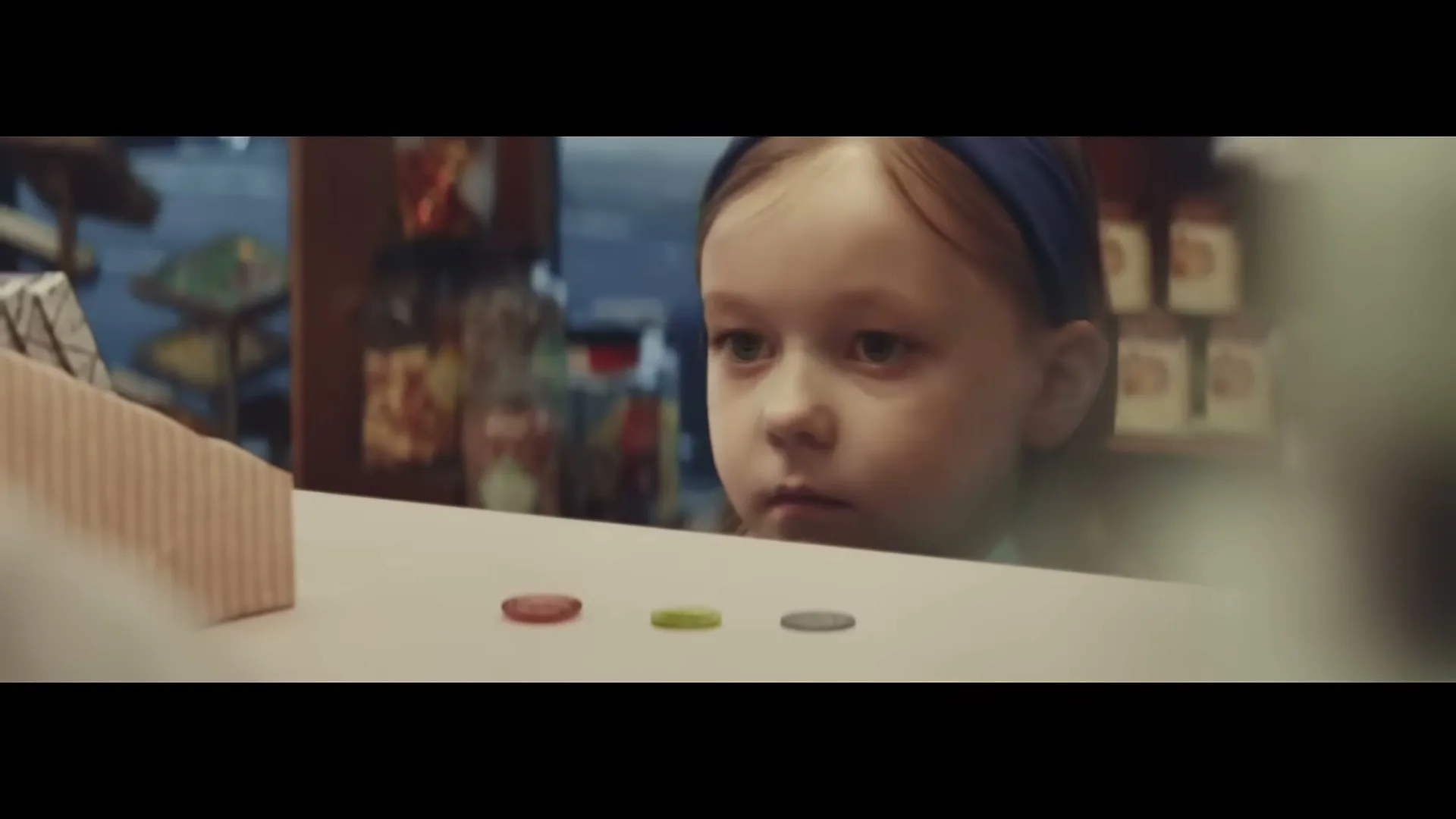 A young girl with a blue headband looks up from behind a counter, with three coins on the counter in front of her.