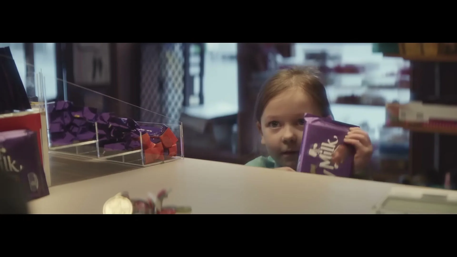 A young girl with light brown hair peeks over a counter, holding a Cadbury Dairy Milk chocolate bar in a store.