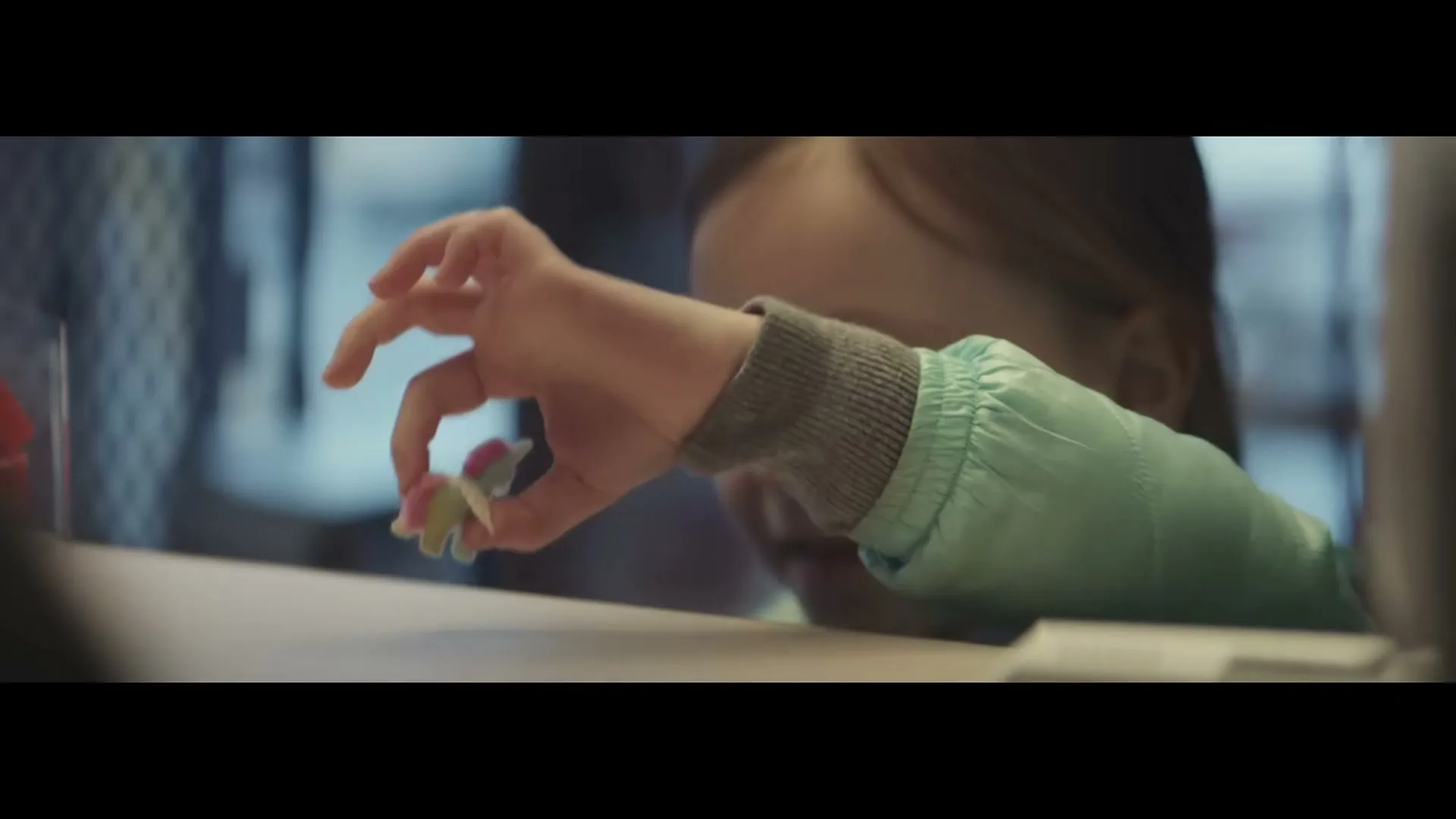 A young girl with brown hair and a light blue jacket plays with a small unicorn toy on a counter, looking up with a happy expression.