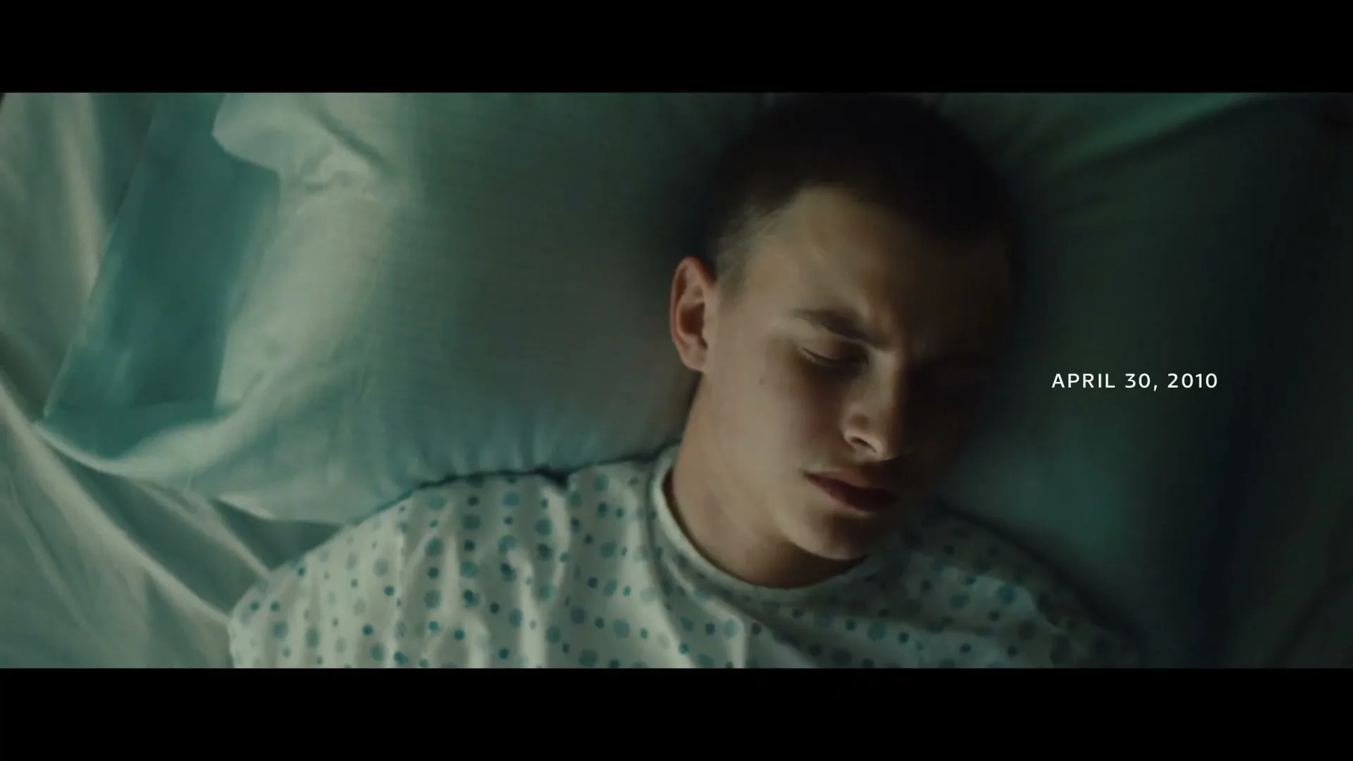 A young male patient with a buzz cut lies in a hospital bed, wearing a gown, with his eyes closed, in a dimly lit room.