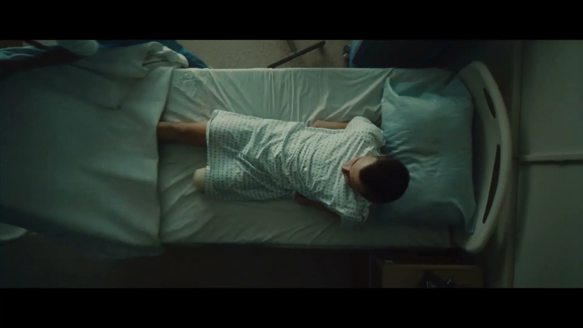 A nurse helps a young male patient in a hospital gown get into bed, pulling the covers over him in a hospital room.