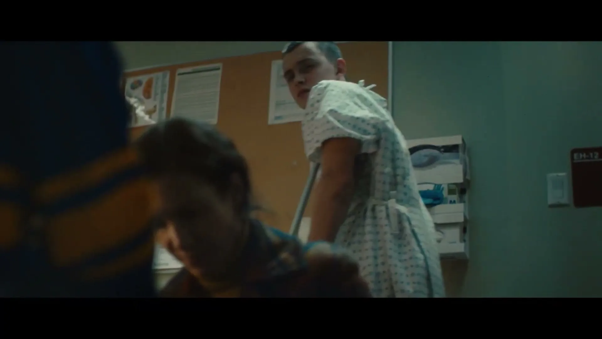 A young woman with dark hair looks down, while a young man in a hospital gown stands behind her, looking at someone off-screen in a hospital room.