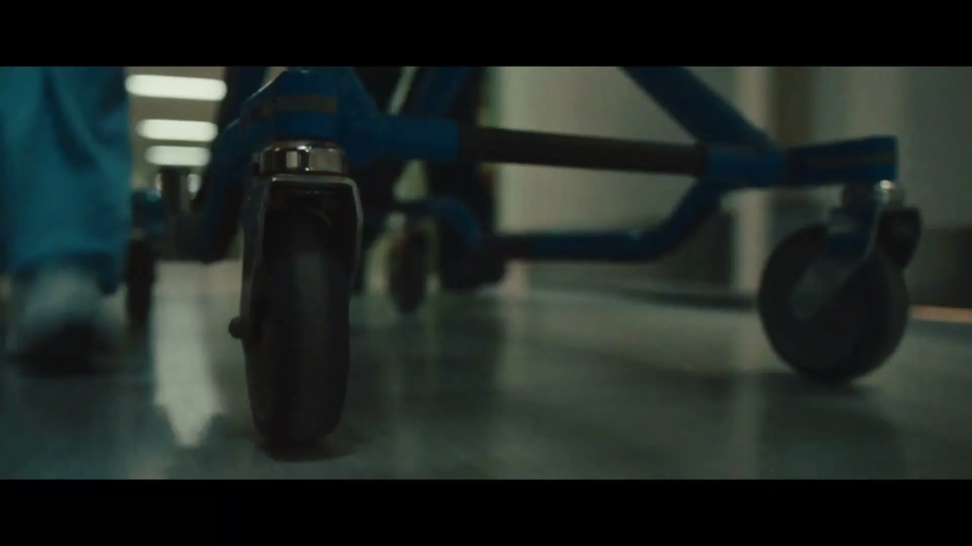 A low-angle shot shows the wheels of a hospital gurney being pushed down a dimly lit hospital hallway.