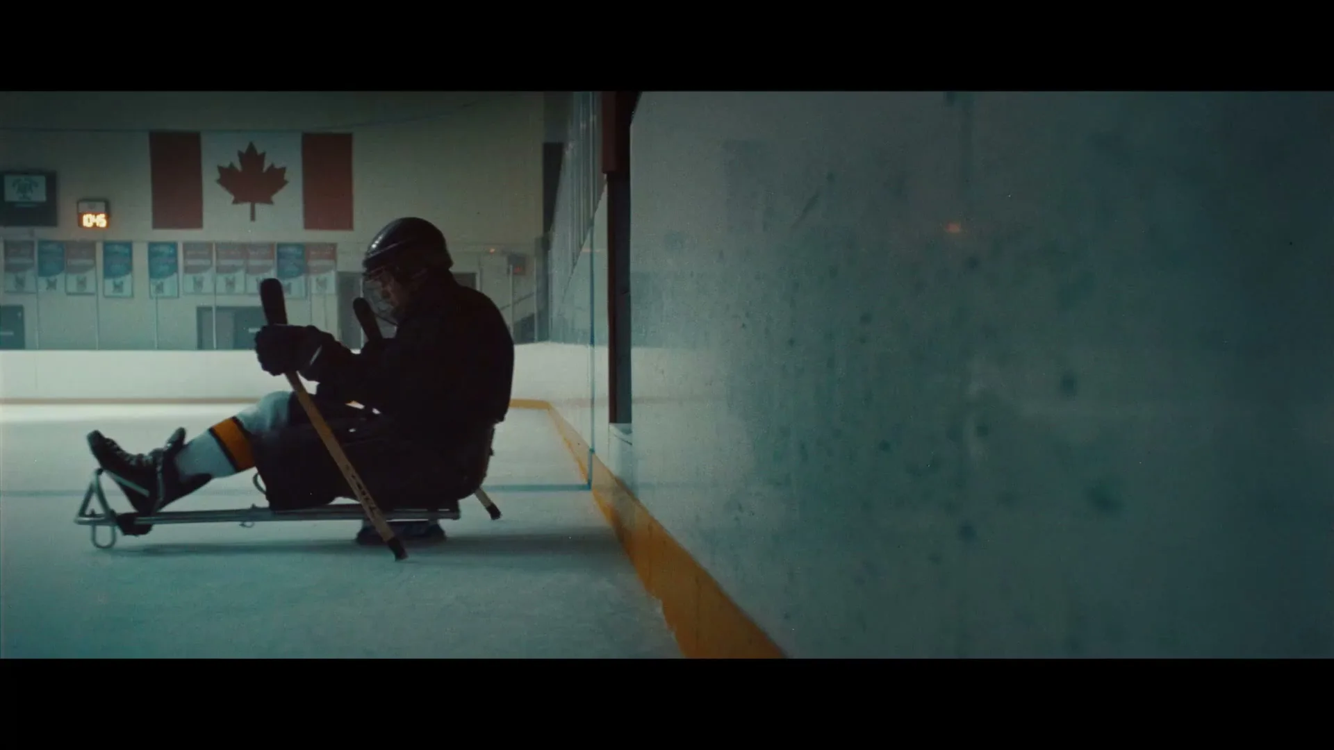 A male para-athlete in hockey gear sits on a sled, holding two hockey sticks, on an ice rink next to the boards, with a Canadian flag in the background.