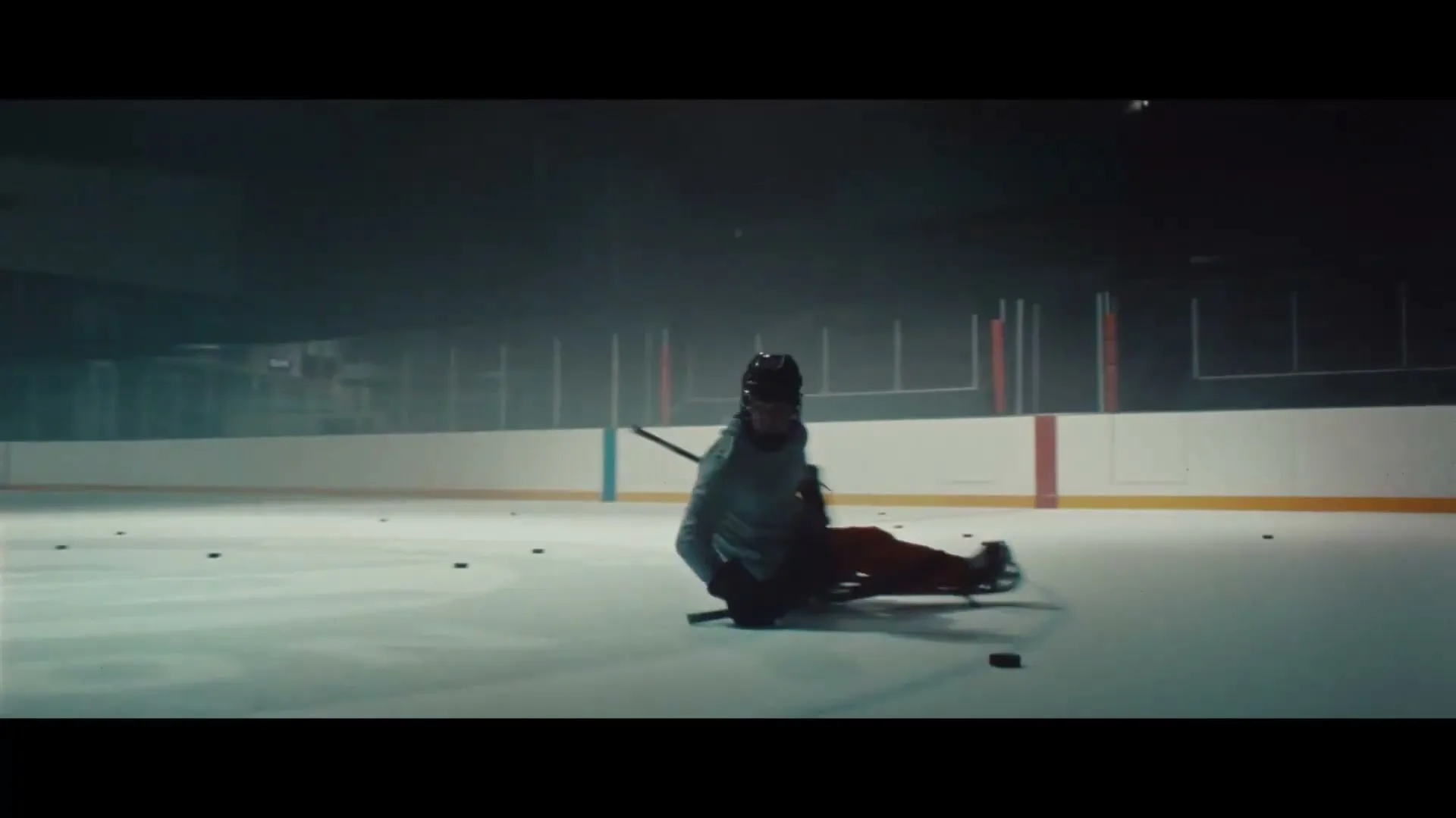 A male para-athlete in a grey hoodie and helmet practices sled hockey on an ice rink, surrounded by scattered pucks, in a dimly lit arena.