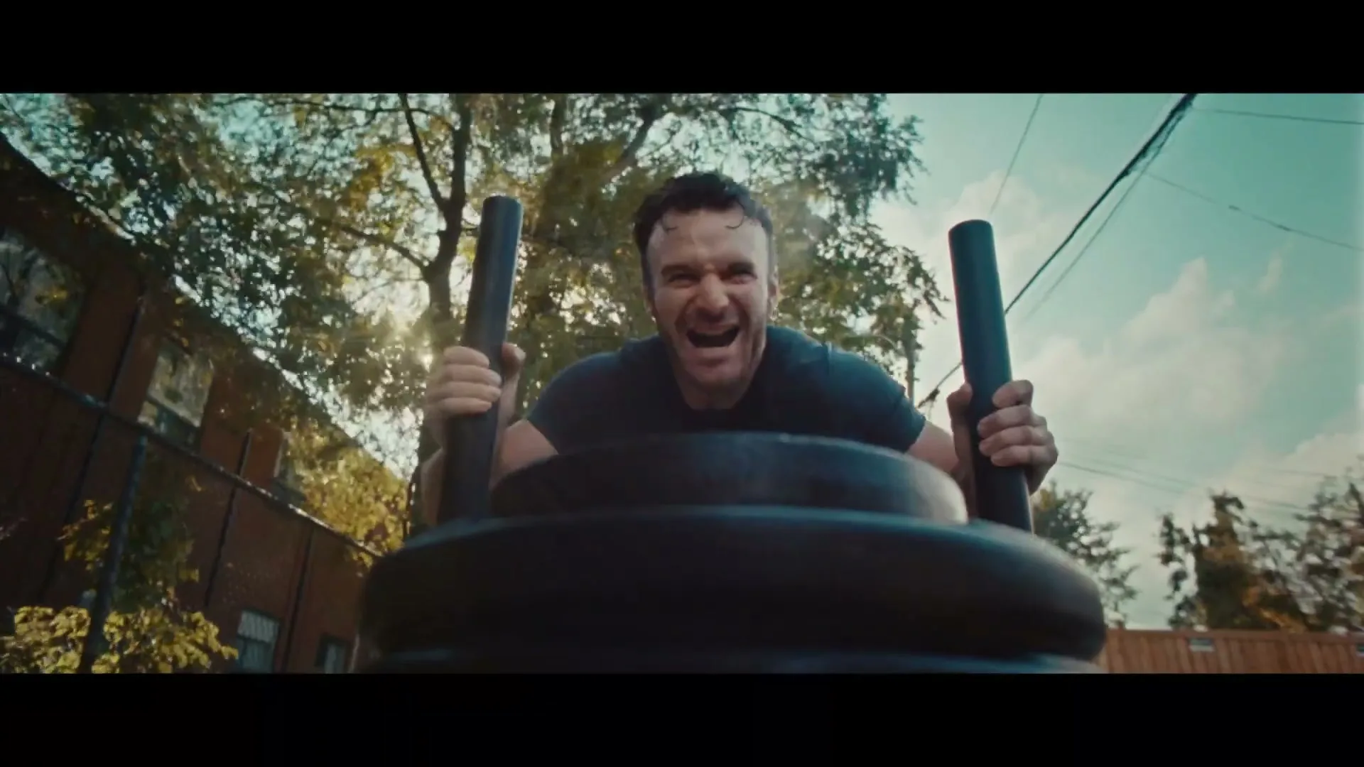 A determined male athlete in his 30s, wearing a black t-shirt, pushes a heavy sled with weights outdoors in an alleyway during the day.