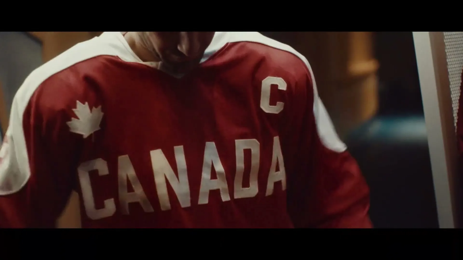 A male hockey player pulls on a red and white Canada jersey in a locker room.