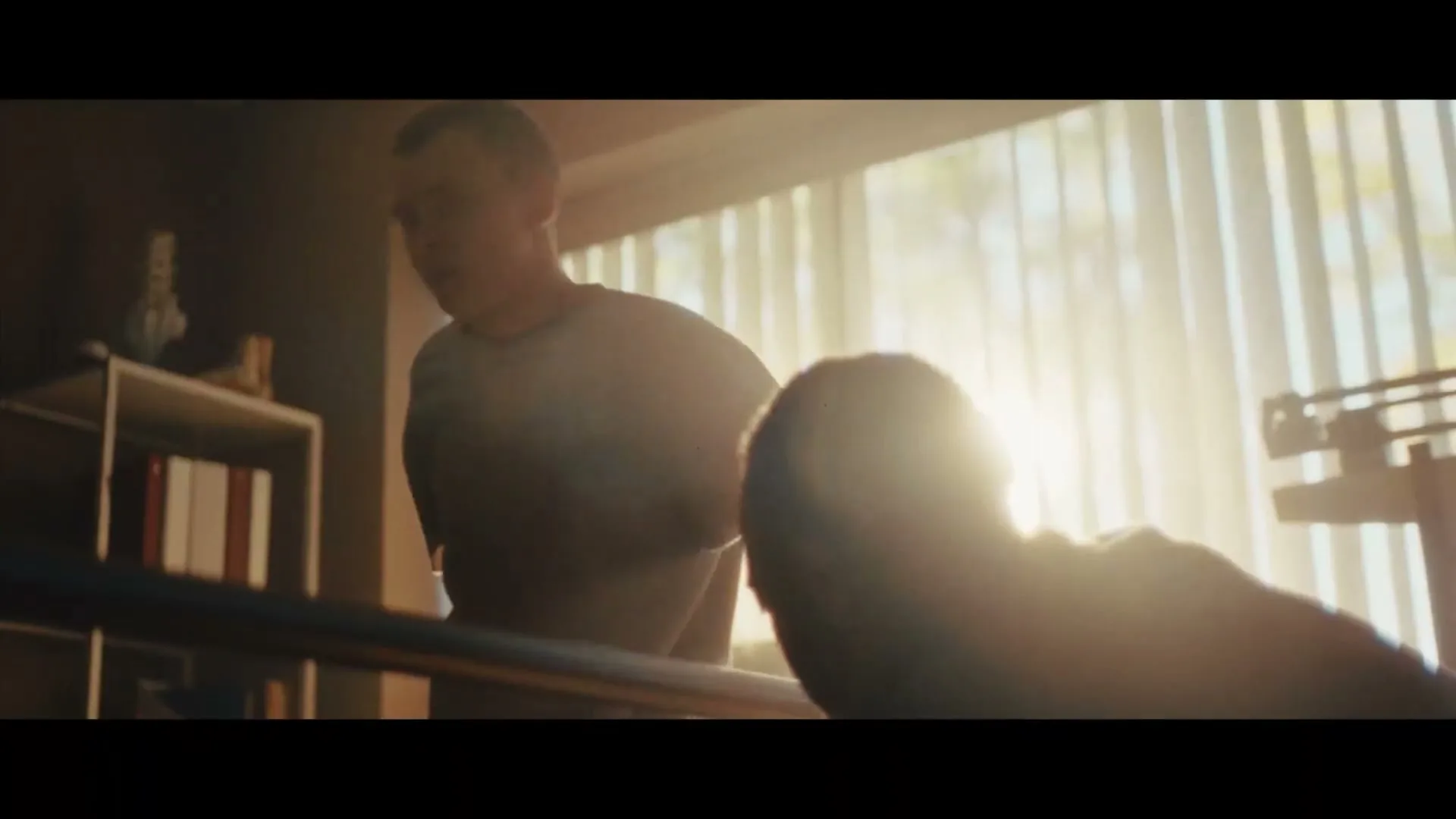 A young man in a grey t-shirt walks with assistance from a physical therapist, silhouetted by bright sunlight streaming through a window in a rehabilitation room.