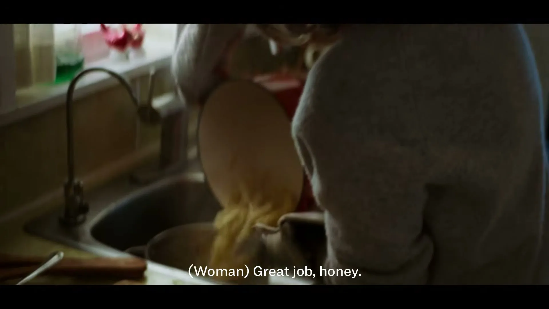 A woman with light brown hair, wearing a white sweater, pours food from a red pot into a sink in a sunlit kitchen.
