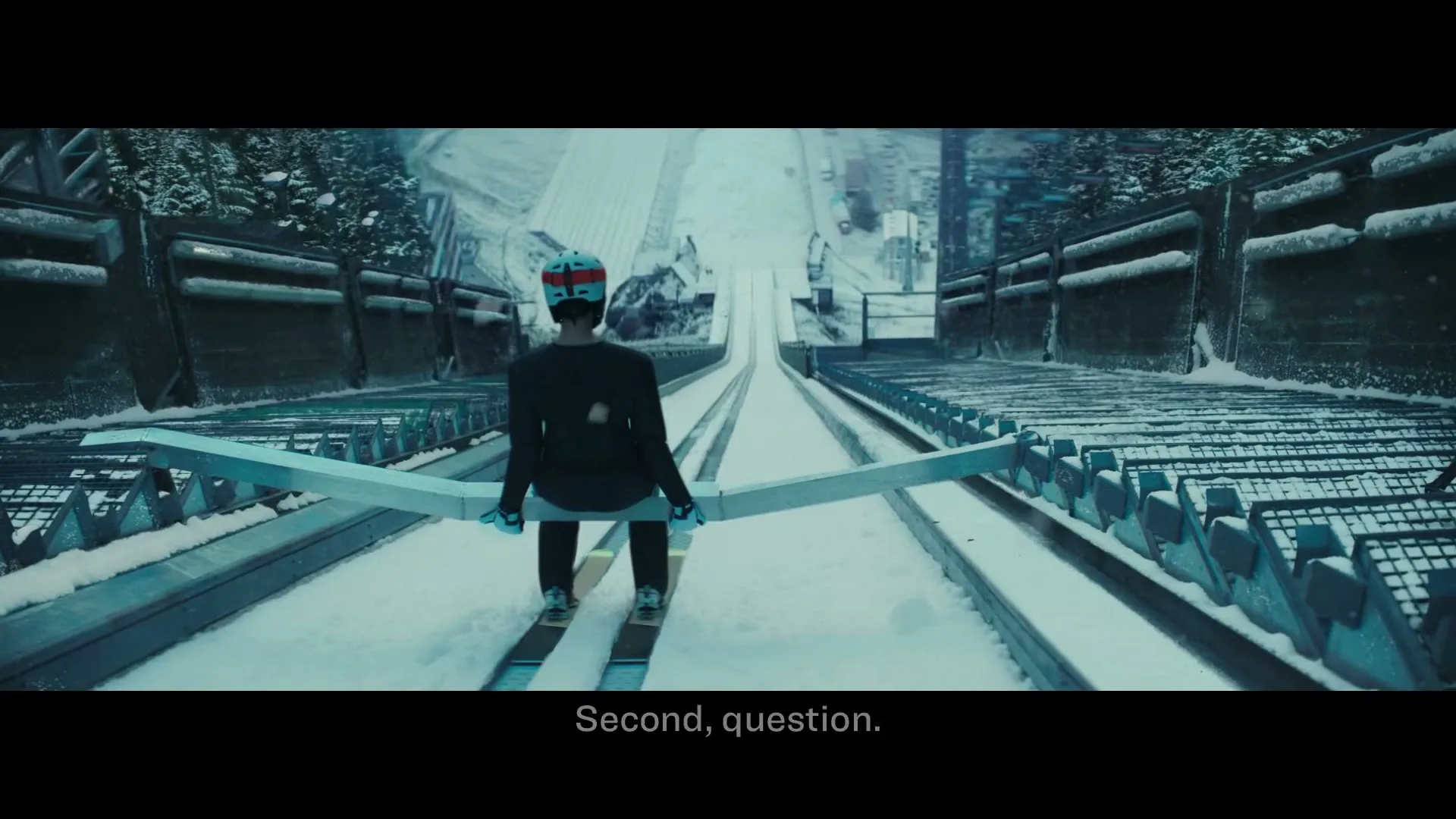 A lone ski jumper in a black suit and helmet sits at the top of a snowy ski jump, looking down the slope on a cold day.