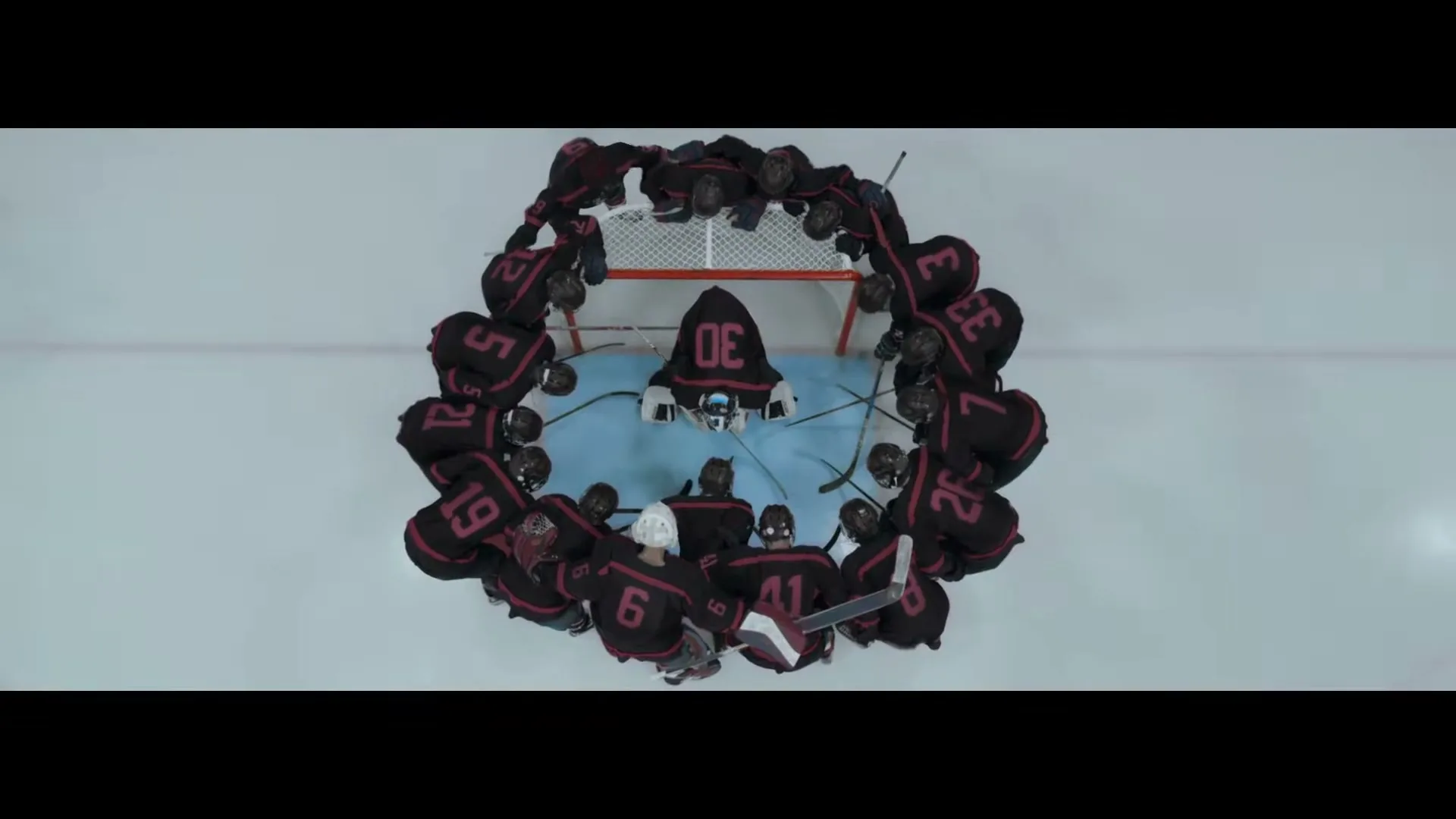 An overhead shot shows a hockey team in dark jerseys huddled in a circle around their goalie on a bright ice rink.