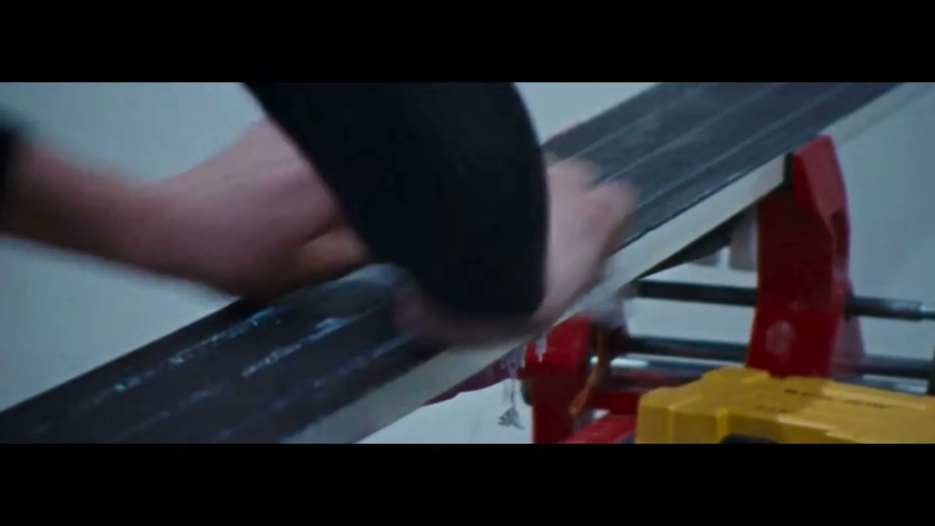 A person's hand scrapes excess wax off a ski base, revealing fresh wax and snow-like shavings, in a close-up shot.