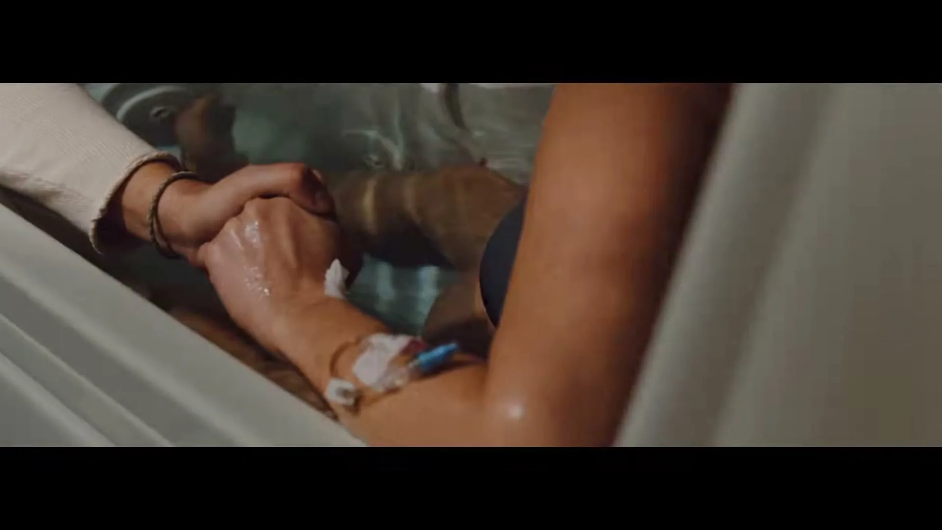 Two people hold hands in a bathtub, one with an IV in their arm, in a close-up shot.
