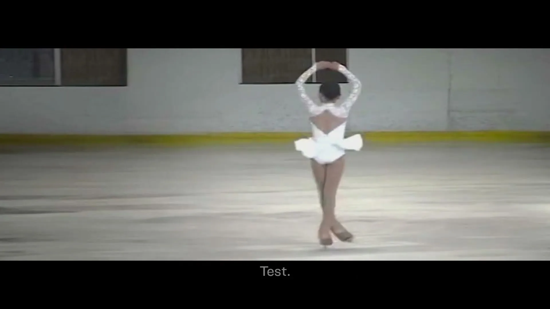 A young female ice skater in a white costume performs a spin on an ice rink, with her arms raised above her head.