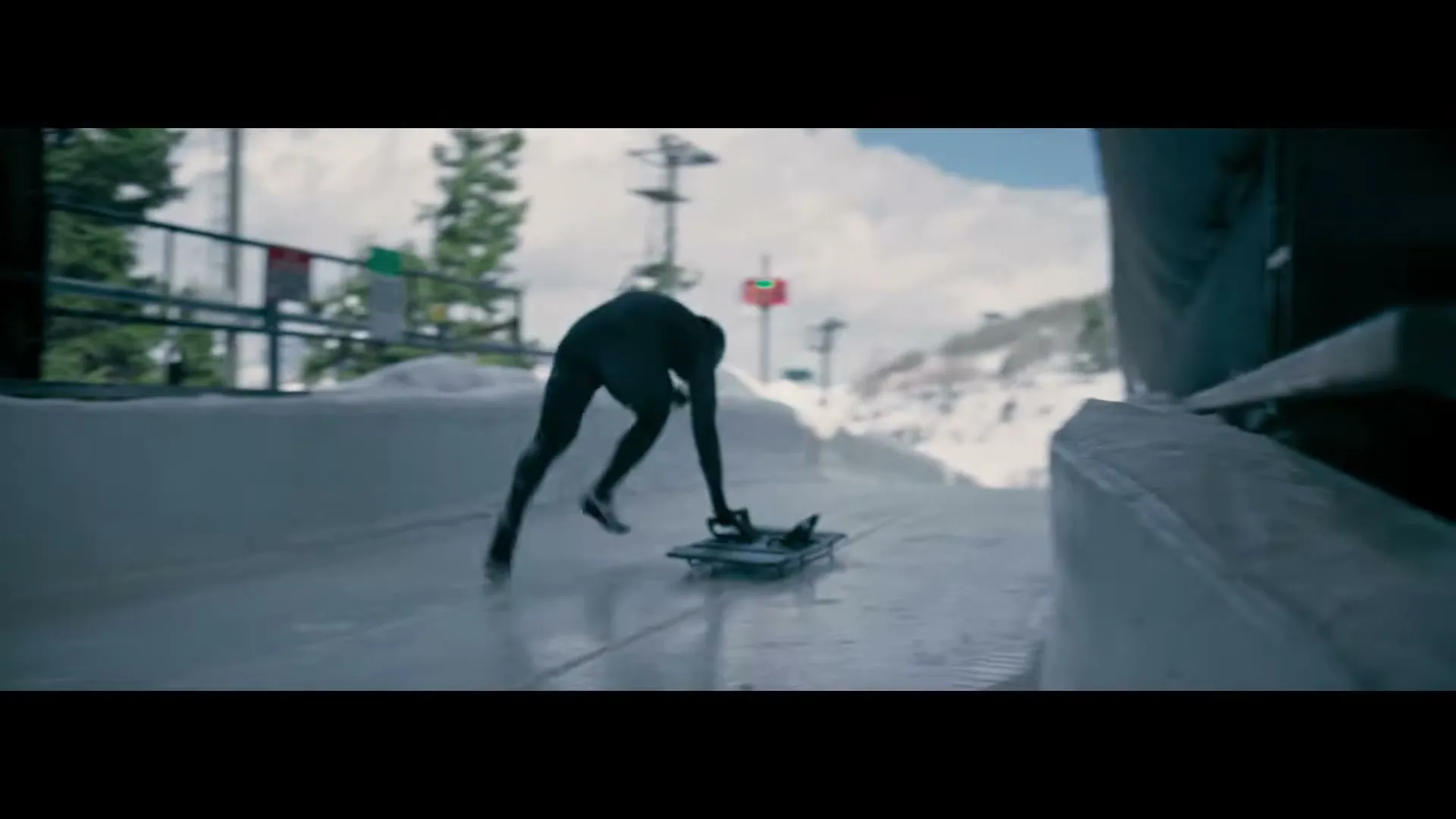 A male athlete in a black speed suit pushes off on a skeleton sled at the start of an ice track on a snowy mountain.