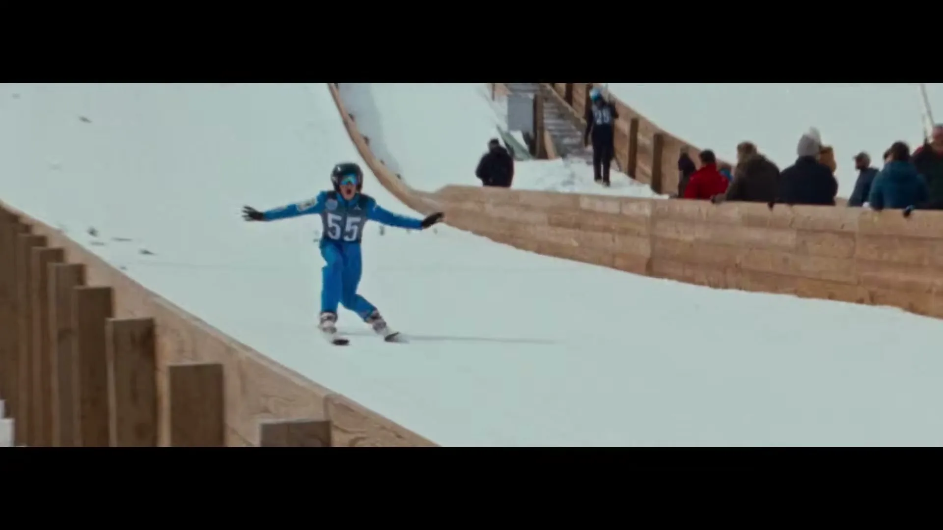 A female skier in a blue suit with number 55 on it, lands on a snowy ski jump slope, with spectators in the background.