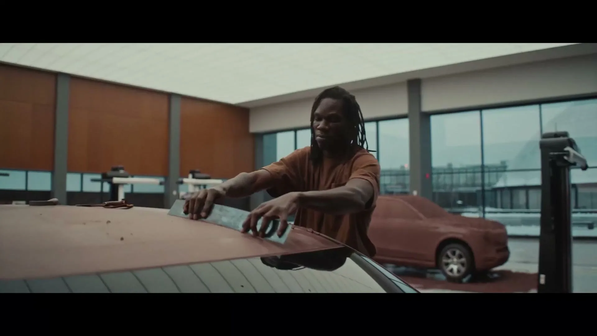 A man with dreadlocks in a brown t-shirt carefully smooths a clay-like substance onto the roof of a car in a modern workshop.