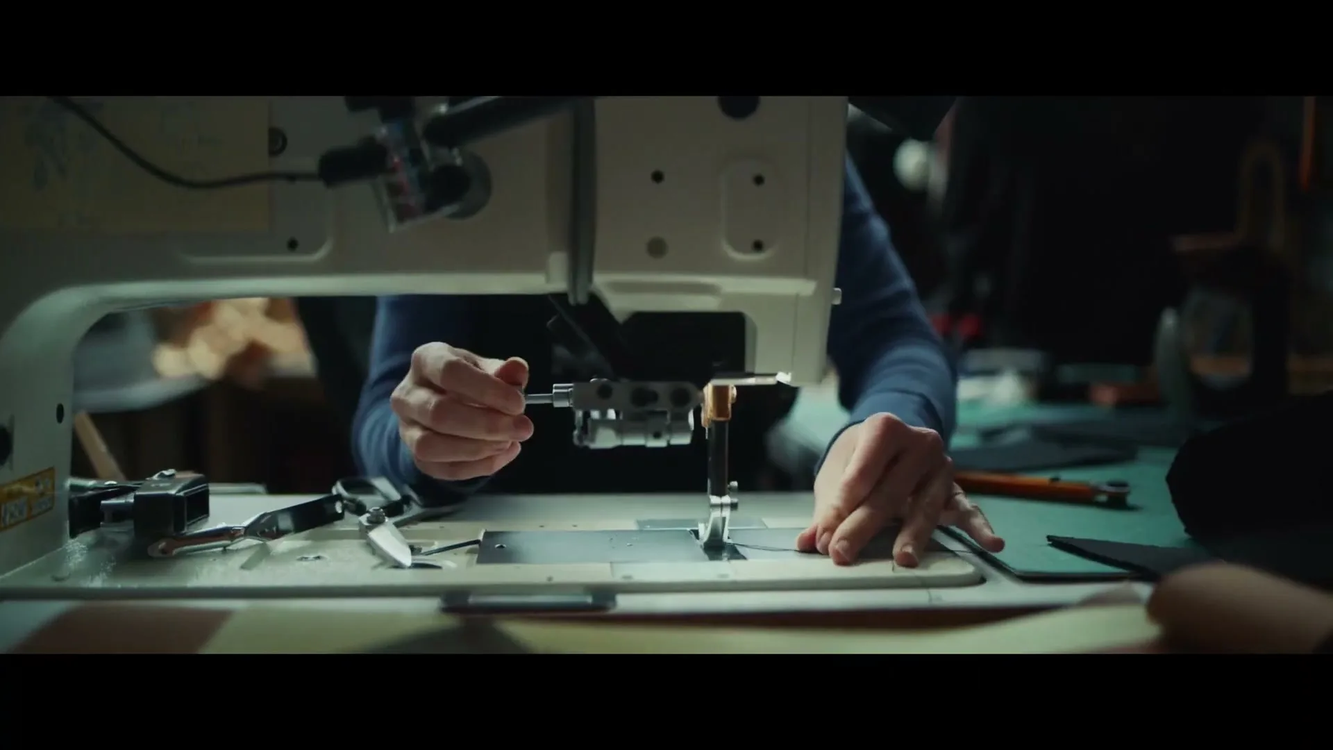 A person's hands guide material through a sewing machine, working on a craft project in a dimly lit workshop.