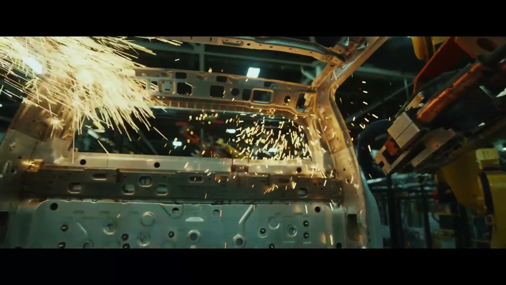 A robotic arm welds the metal frame of a car, creating a shower of sparks in a dimly lit factory.