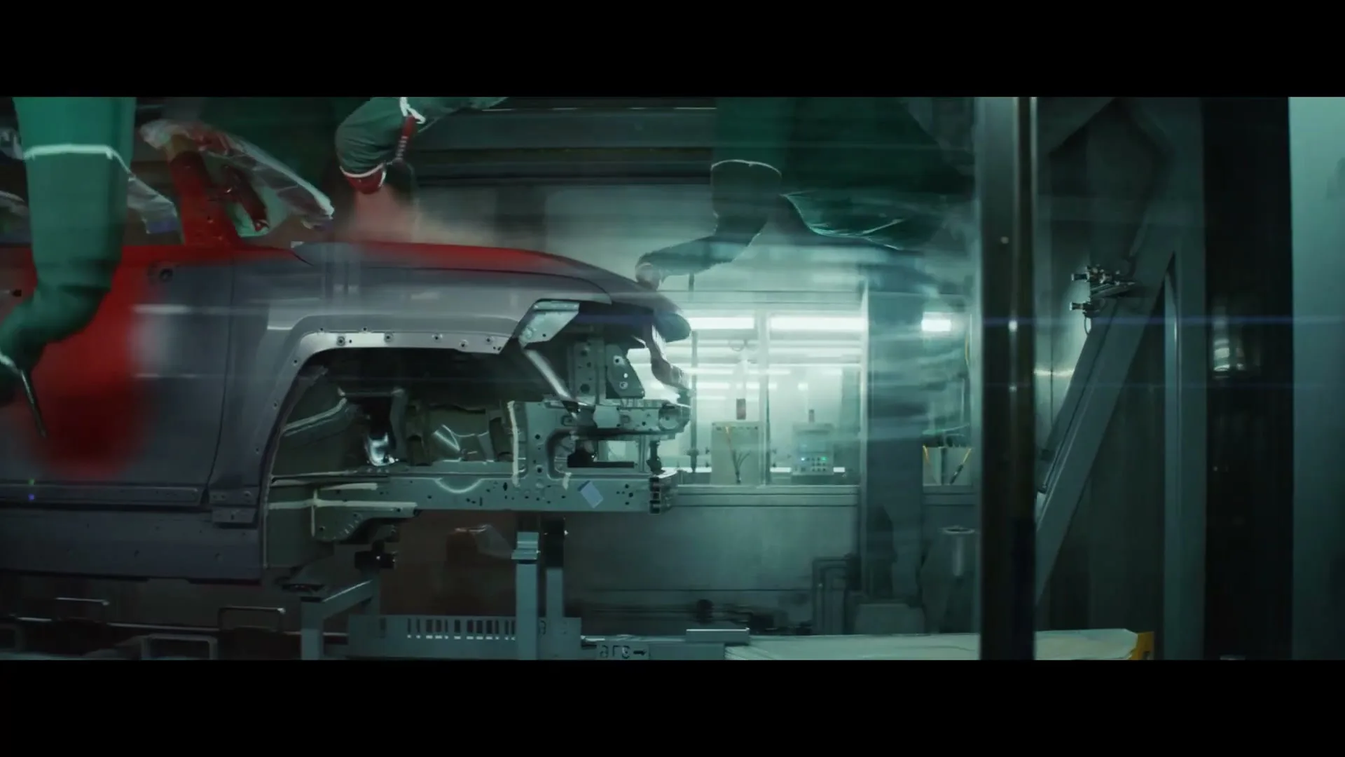 A robotic arm sprays red paint onto the front chassis of a car in a dimly lit factory setting, showcasing the manufacturing process.