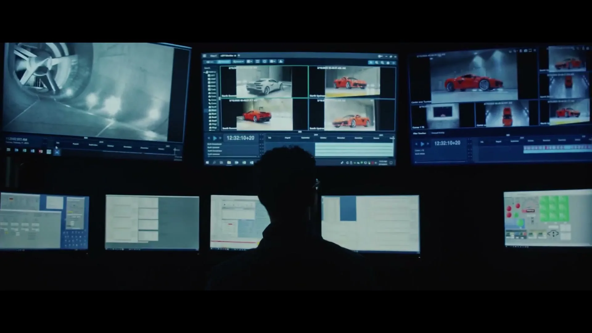 A man in silhouette watches multiple computer screens displaying car designs and data in a dark control room.