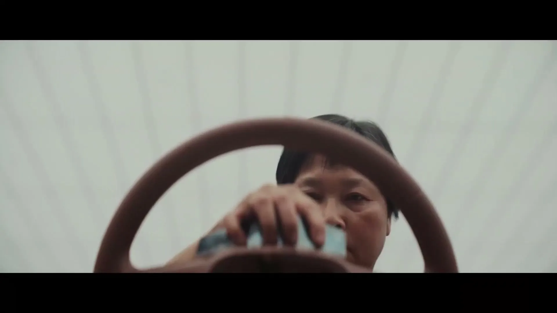 A middle-aged person with short dark hair is seen from a low angle, looking over a steering wheel, holding a small blue object.