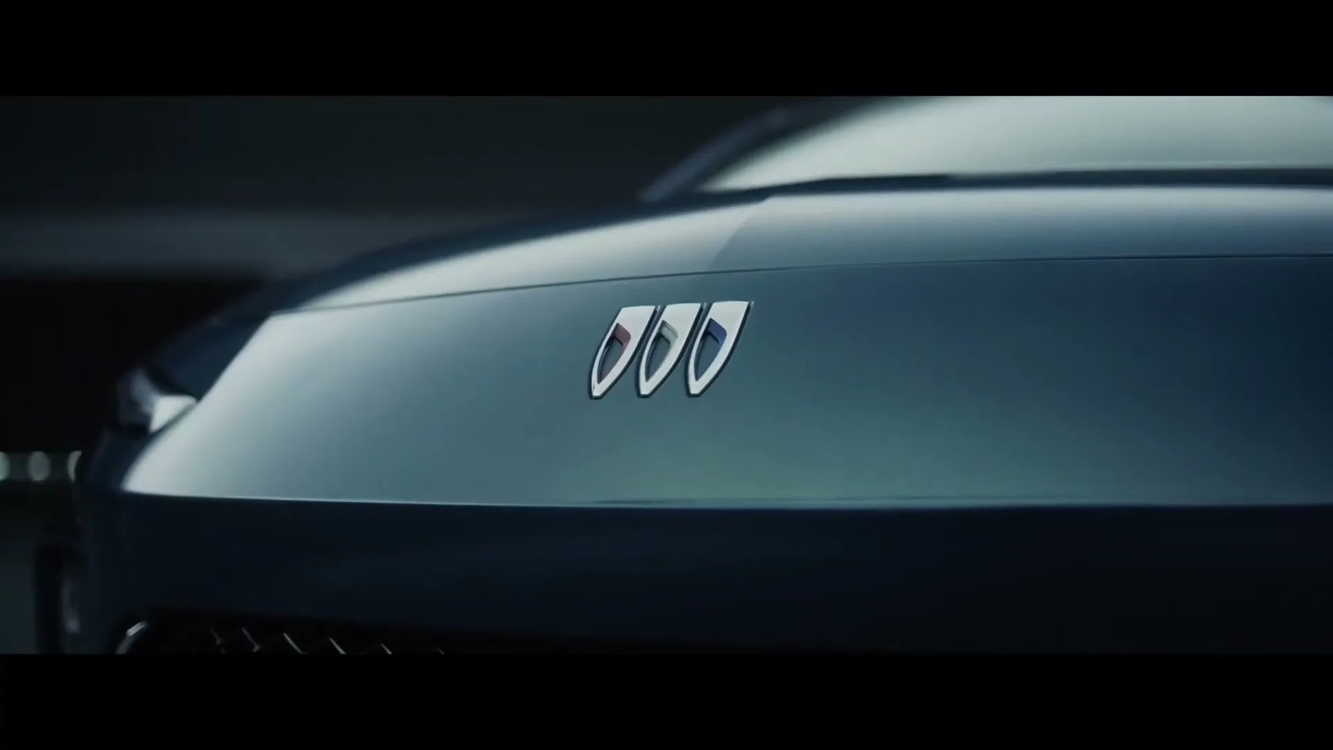 A close-up shot of a dark blue car's hood, featuring a prominent silver emblem with three distinct shapes, in a dimly lit setting.