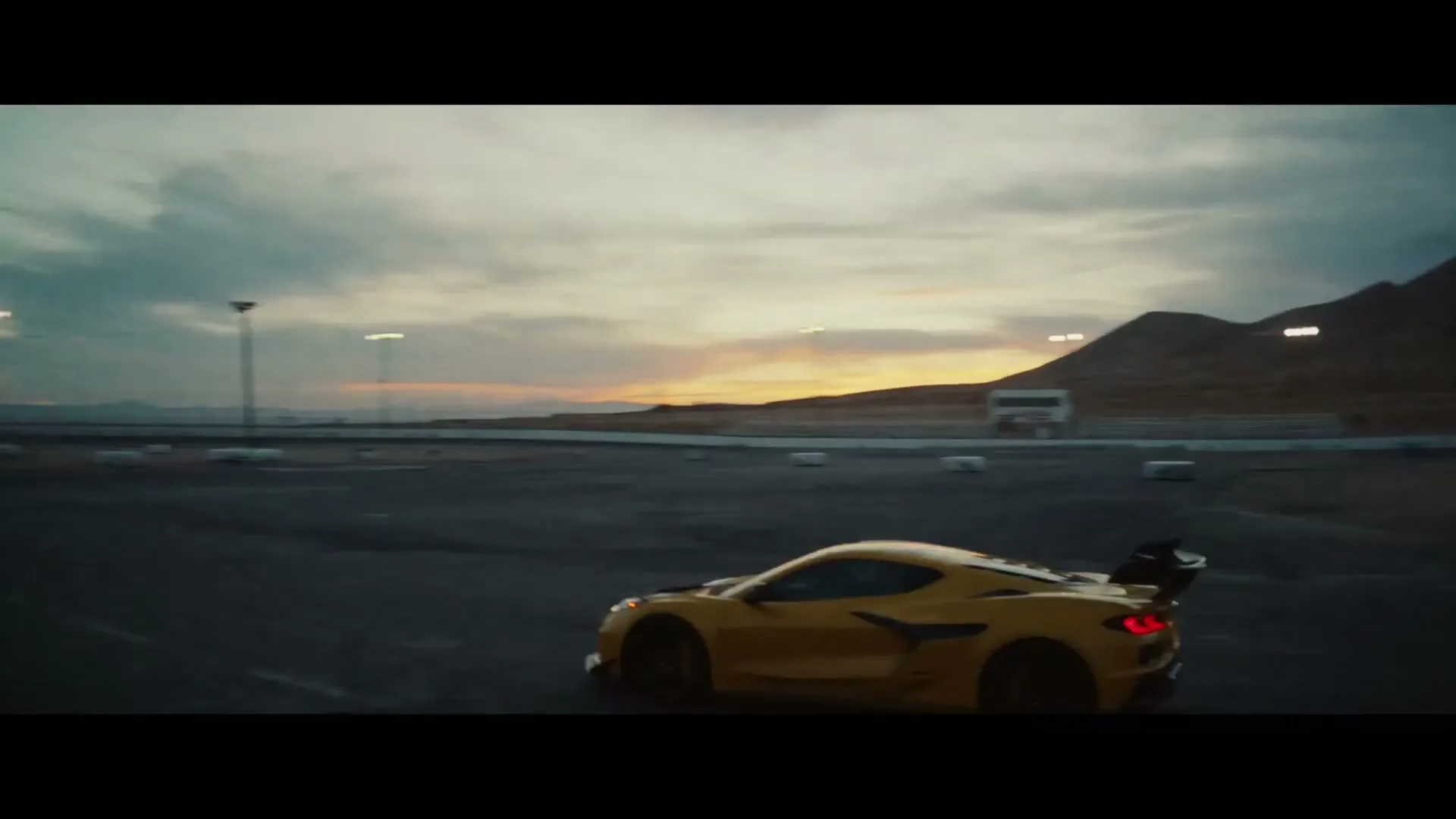 A yellow sports car speeds around a race track at dusk, with mountains and a cloudy sky in the background.