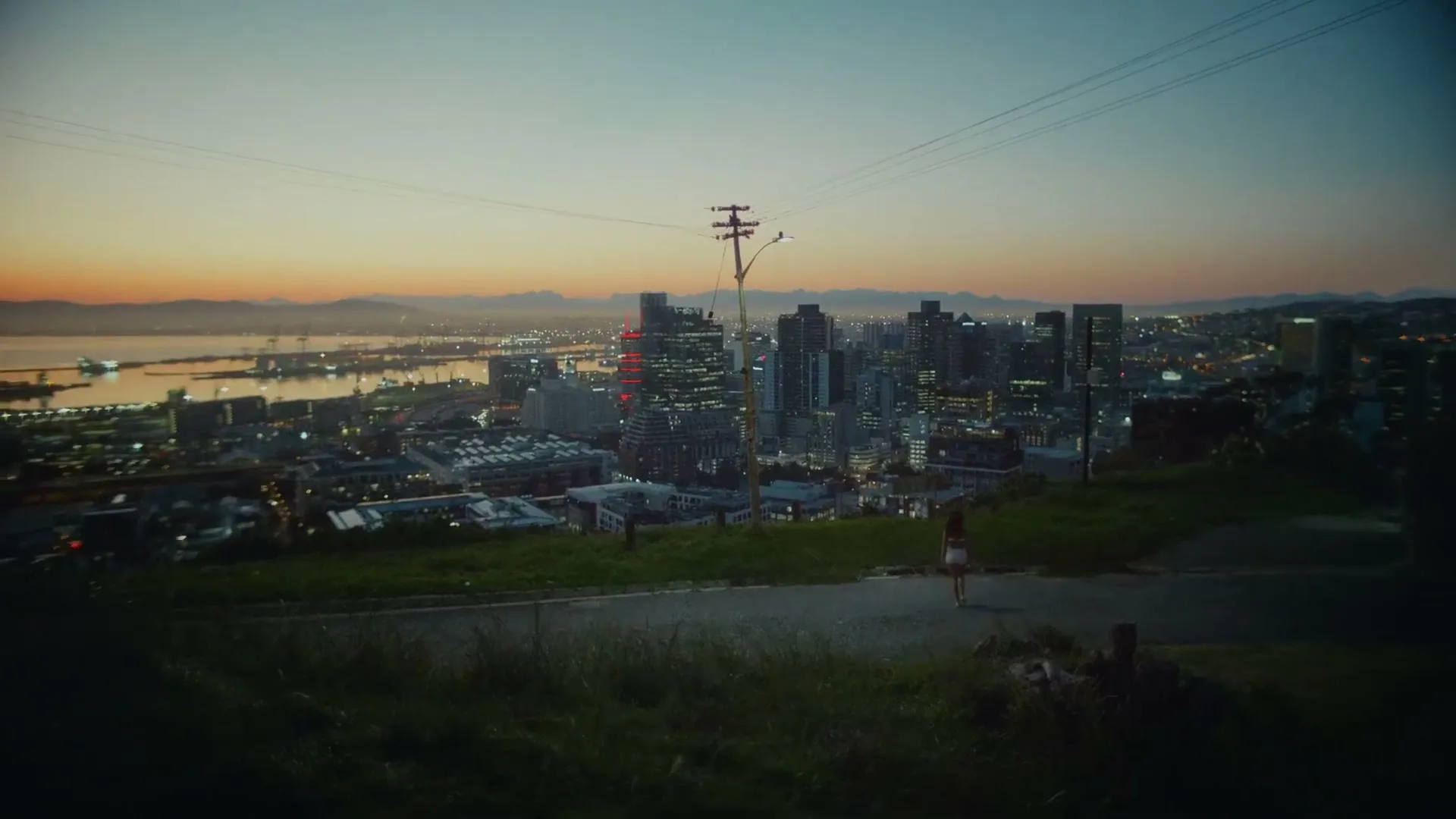 A young woman in a white dress walks down a hill overlooking a city skyline and harbor at dusk.