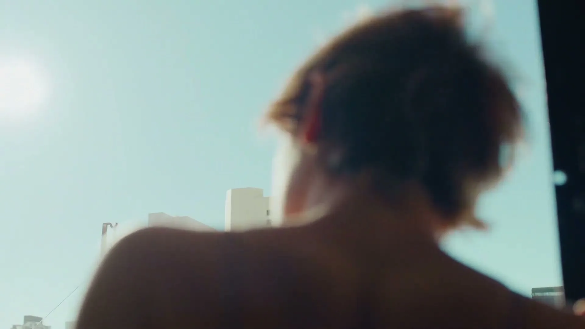 A blurry shot of a person's back and shoulder, looking out a window at a bright blue sky and distant buildings.
