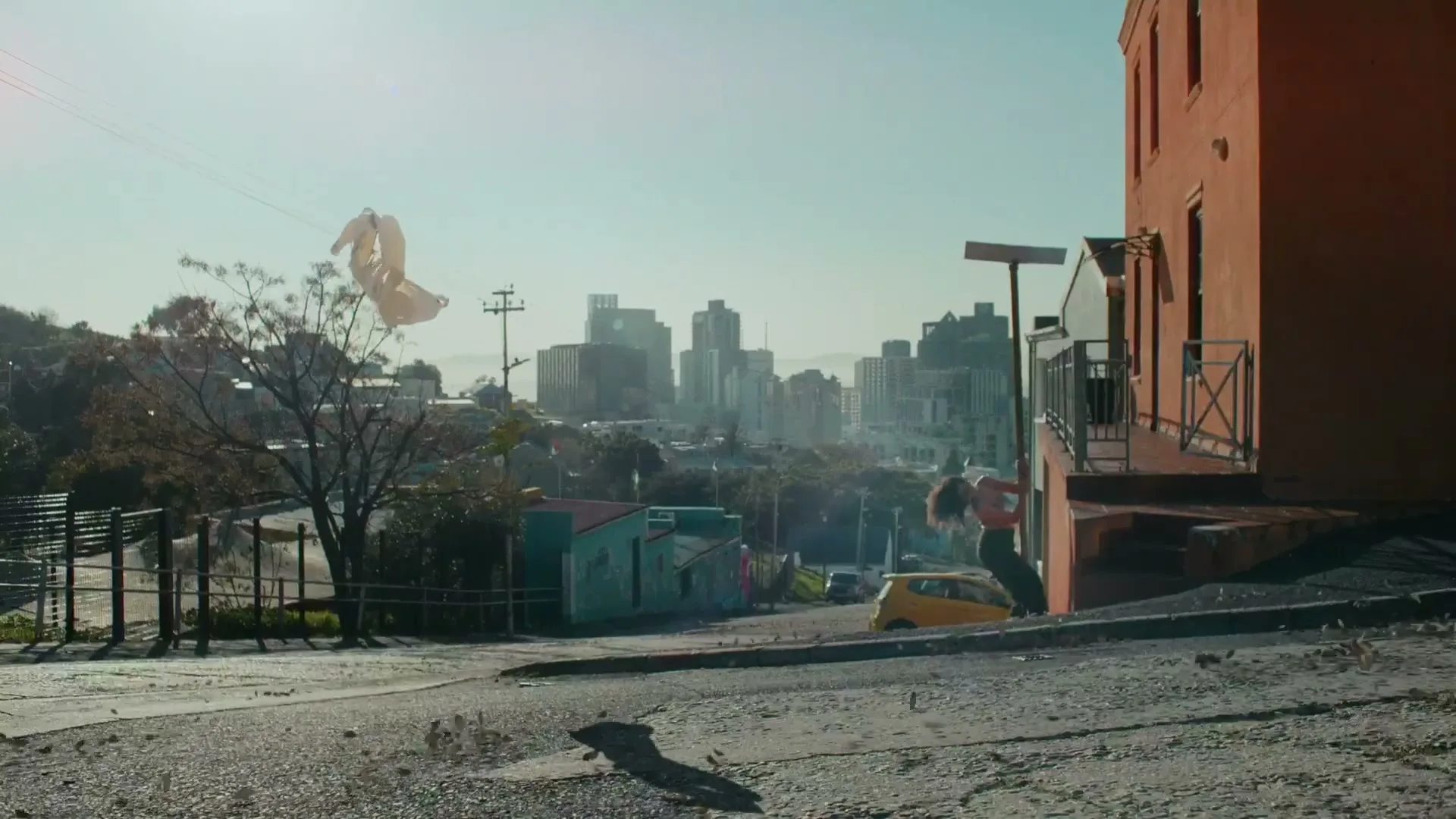A young woman in a sports bra and shorts swings around a pole on a sunny day in a city with a skyline in the background.